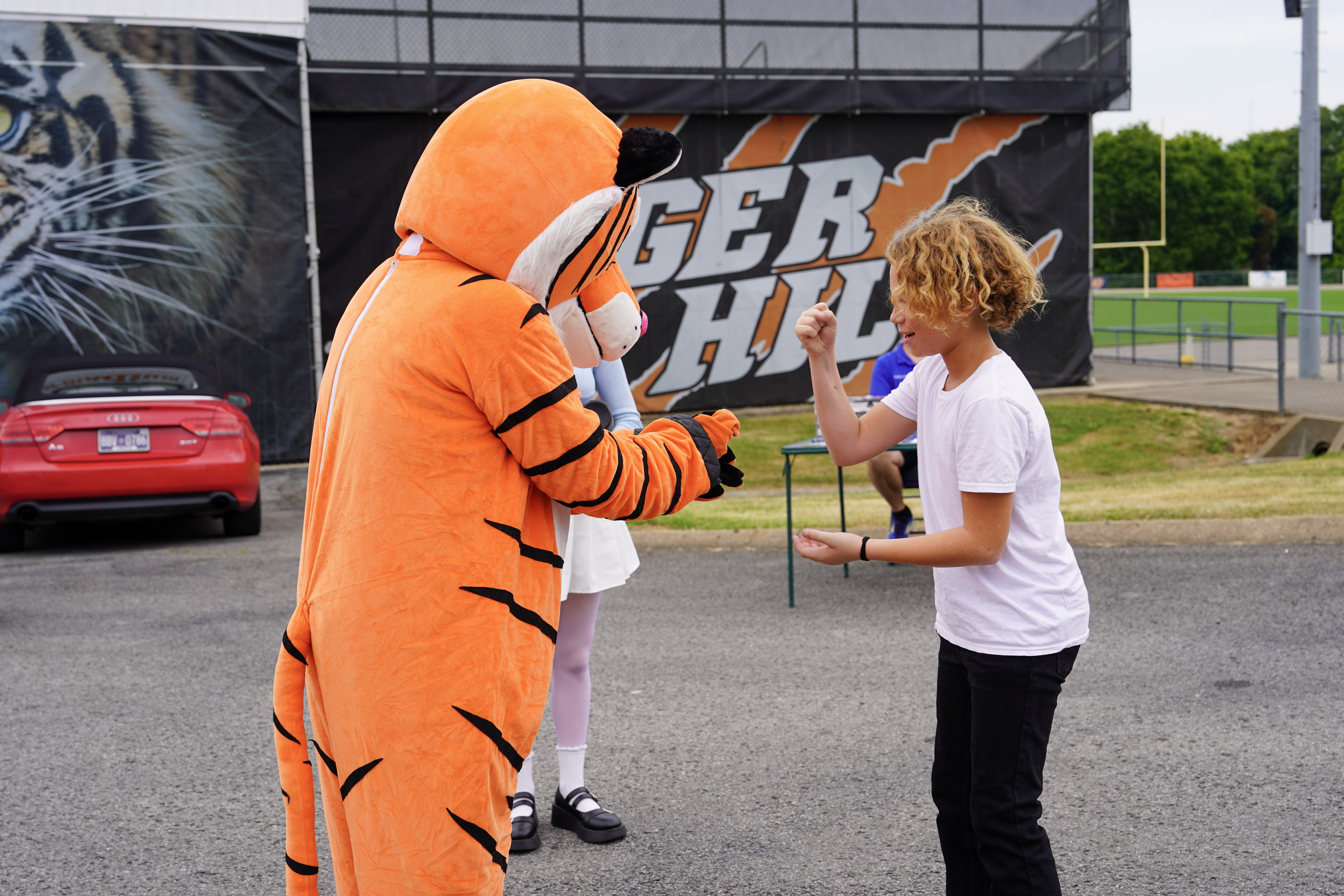 A person in a tiger mascot costume plays rock-paper-scissors with a smiling child wearing a white t-shirt. A girl in white stands in the background near a Tiger Pride banner and a parked car.