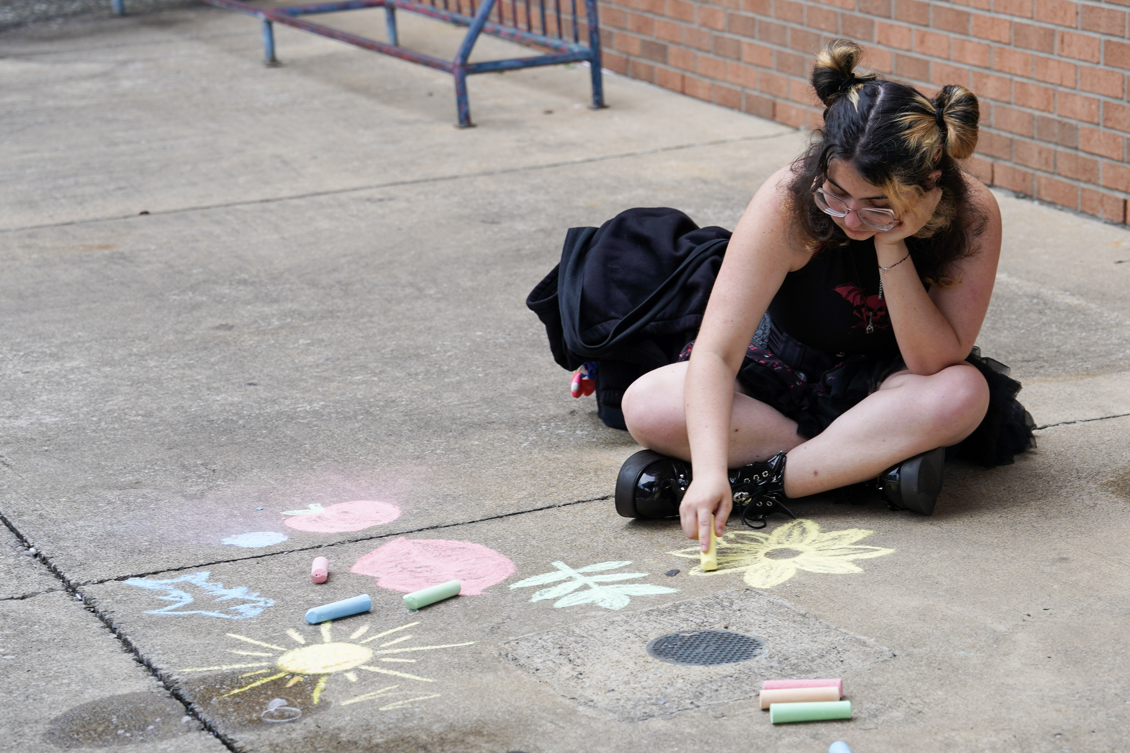 A person with glasses and double buns sits on the ground, drawing colorful chalk art including flowers and suns on a concrete surface. Chalk sticks are scattered nearby, and a brick wall is in the background.
