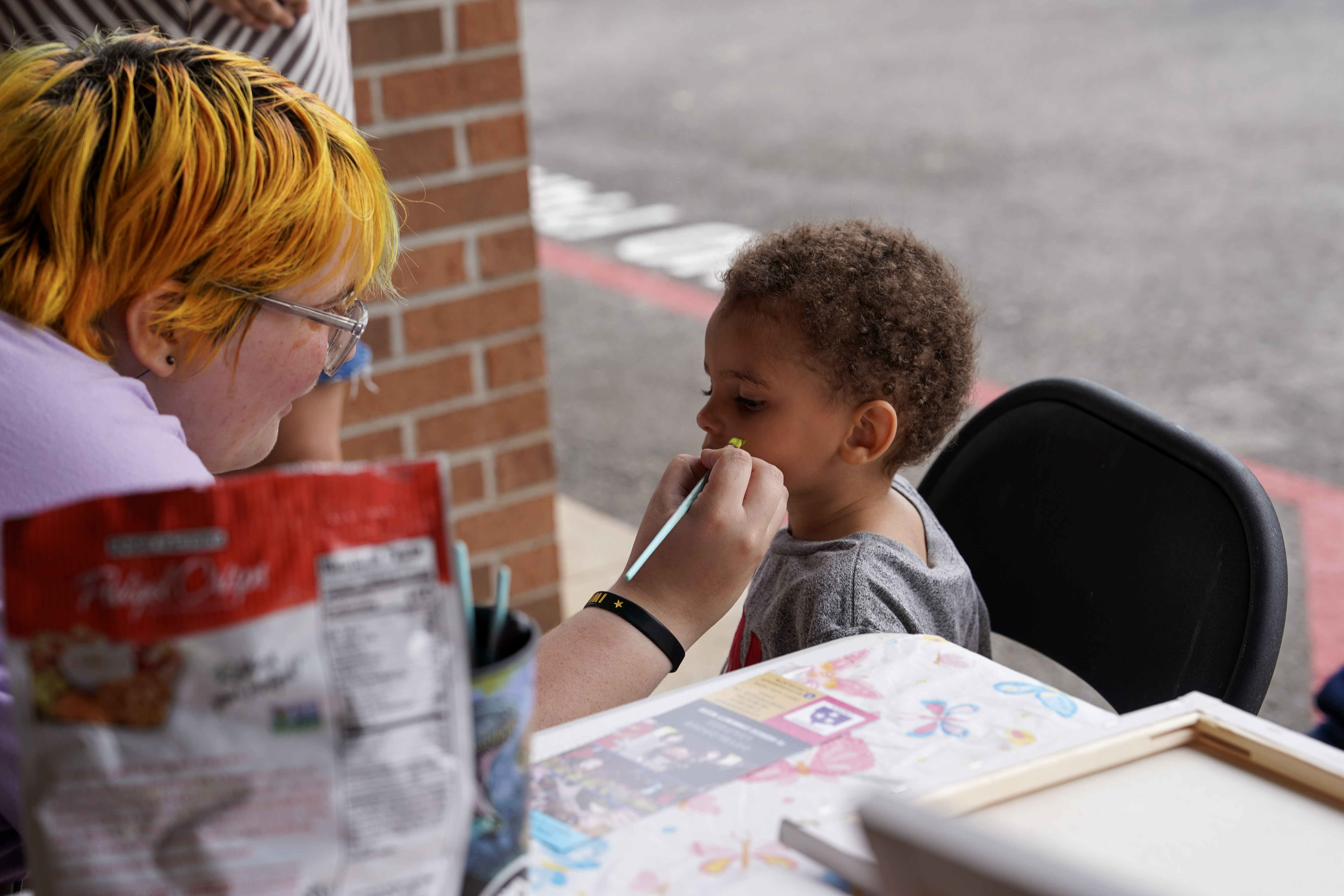 An adult with yellow and orange hair paints a young childs face at an outdoor table covered with art supplies and snacks. The child sits patiently, watching the artist. A brick wall and parking lot are visible in the background.