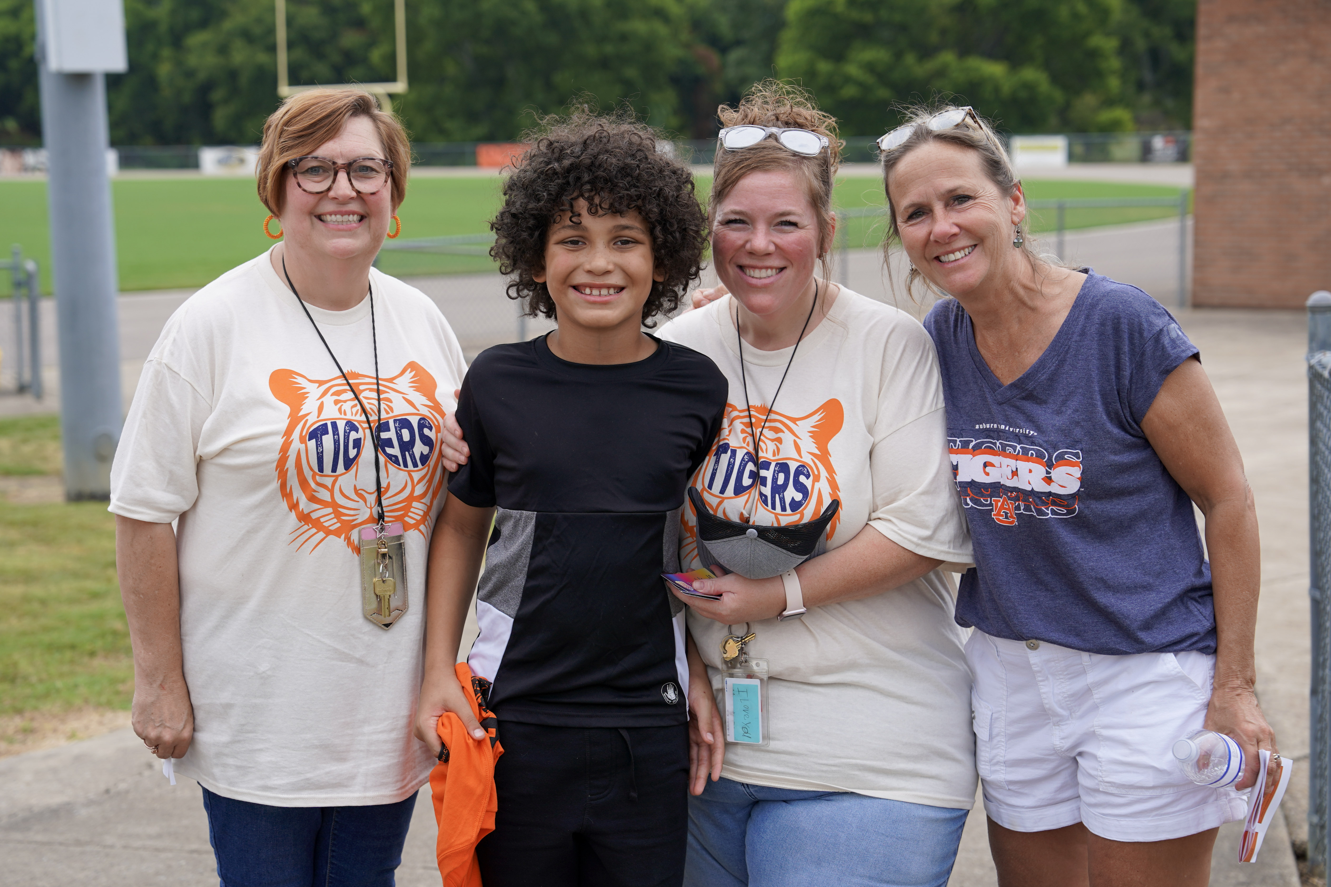 Four smiling adults and one child stand outdoors by a sports field. The adults wear Tigers shirts and lanyards, suggesting school staff or coaches. The child stands in front, and everyone appears happy and relaxed.