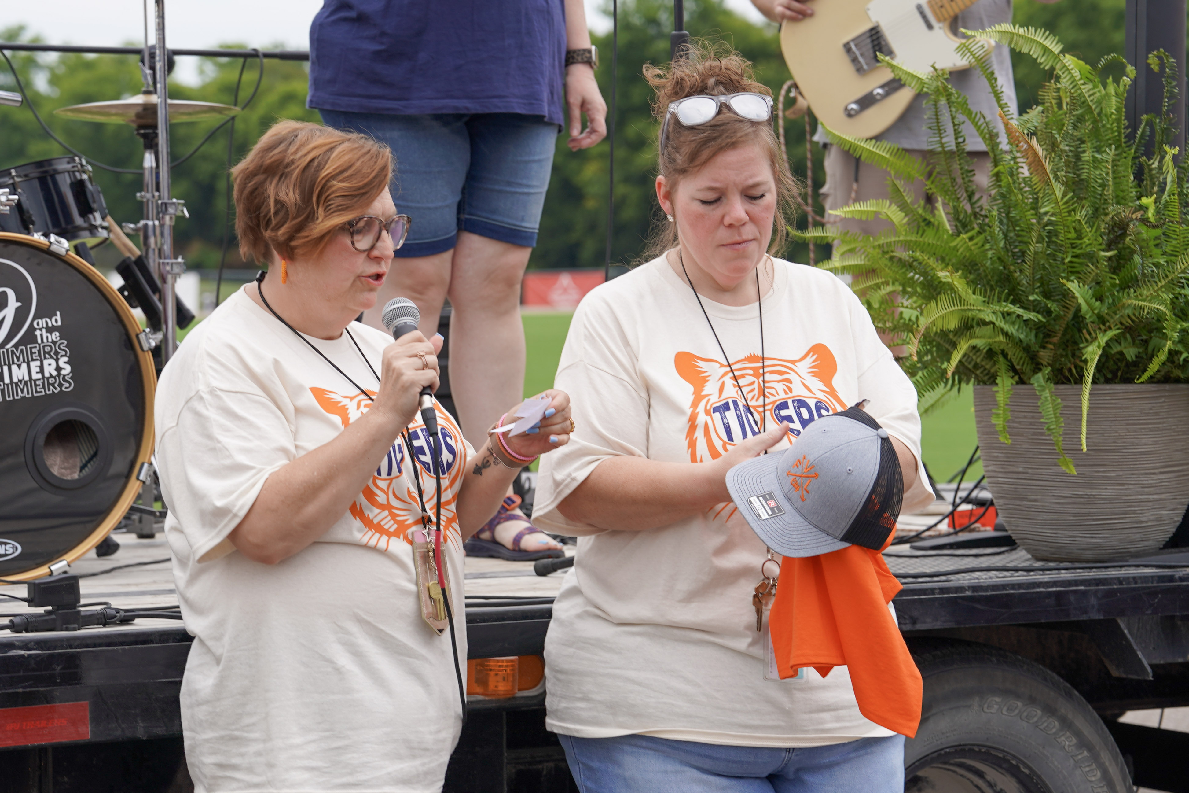 Two women in matching Tigers shirts stand on an outdoor stage; one speaks into a microphone while the other holds a hat and an orange shirt. A drum set, guitar, and potted plant are visible in the background.