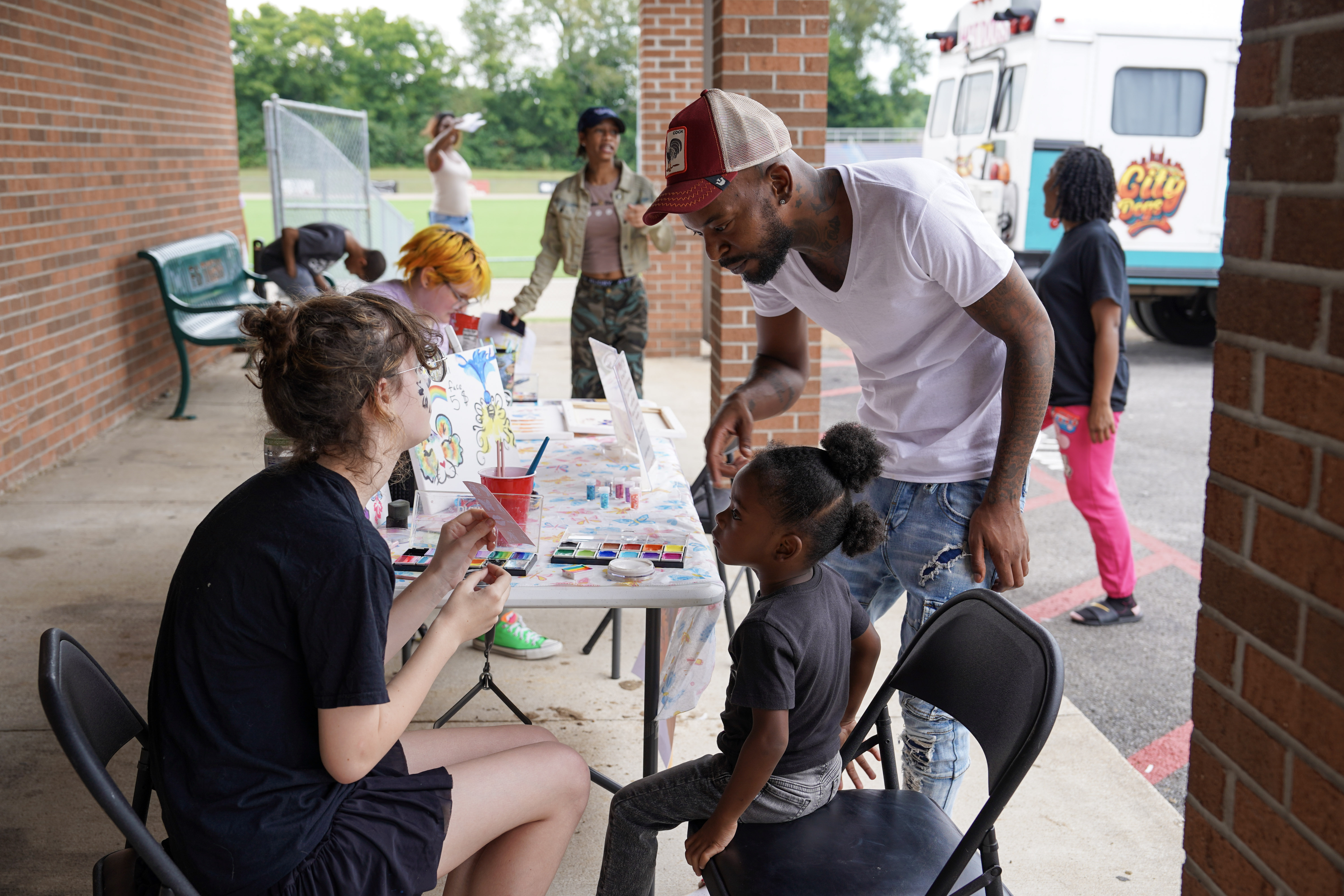 A woman sitting at a table paints a young girls face while a man leans in, speaking to the child. Art supplies are on the table, and other people are nearby outdoors on a covered sidewalk.