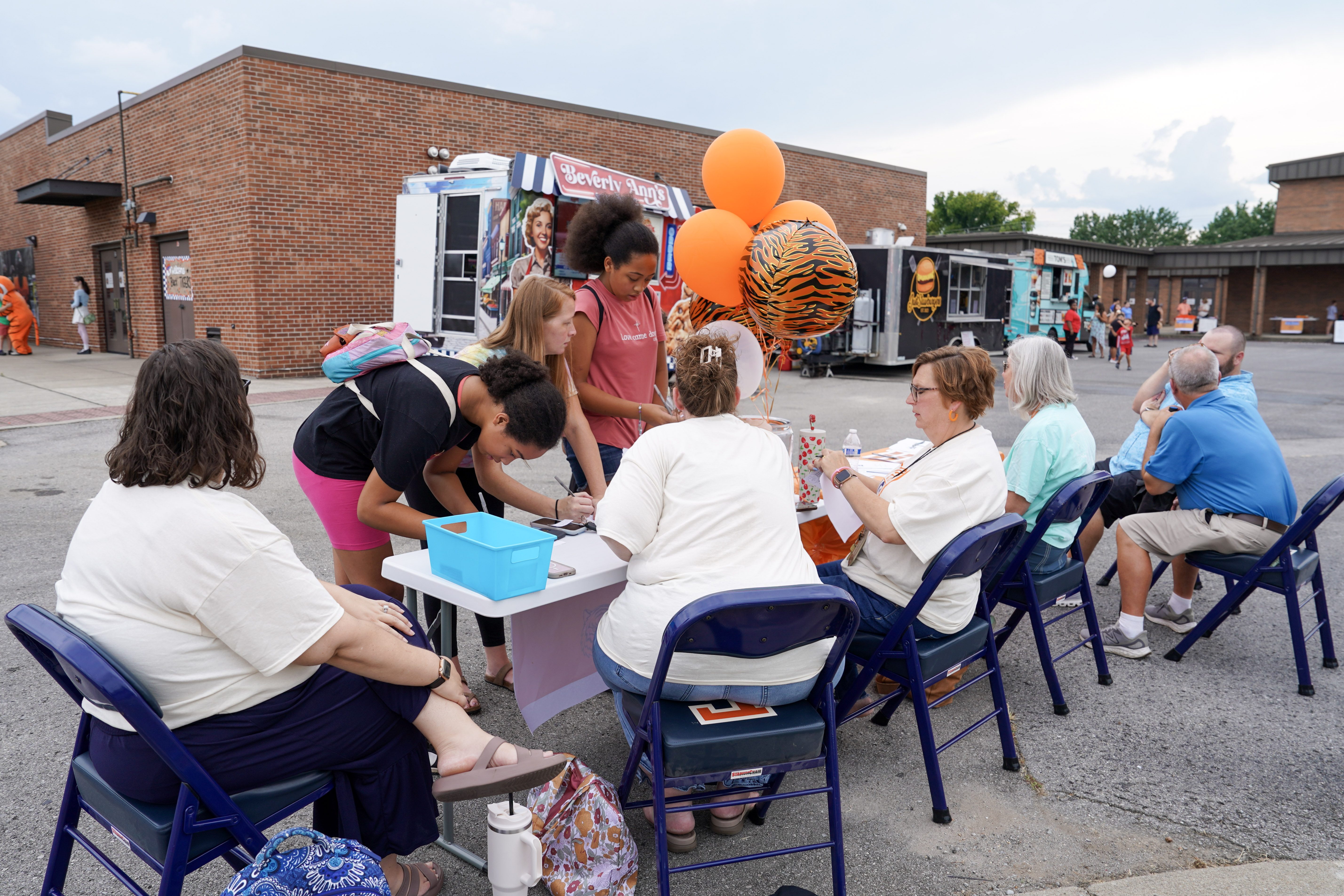 People gather at an outdoor event near food trucks and brick buildings. Several people sit at a table with papers and orange balloons, while others stand and interact with them on a sunny day.