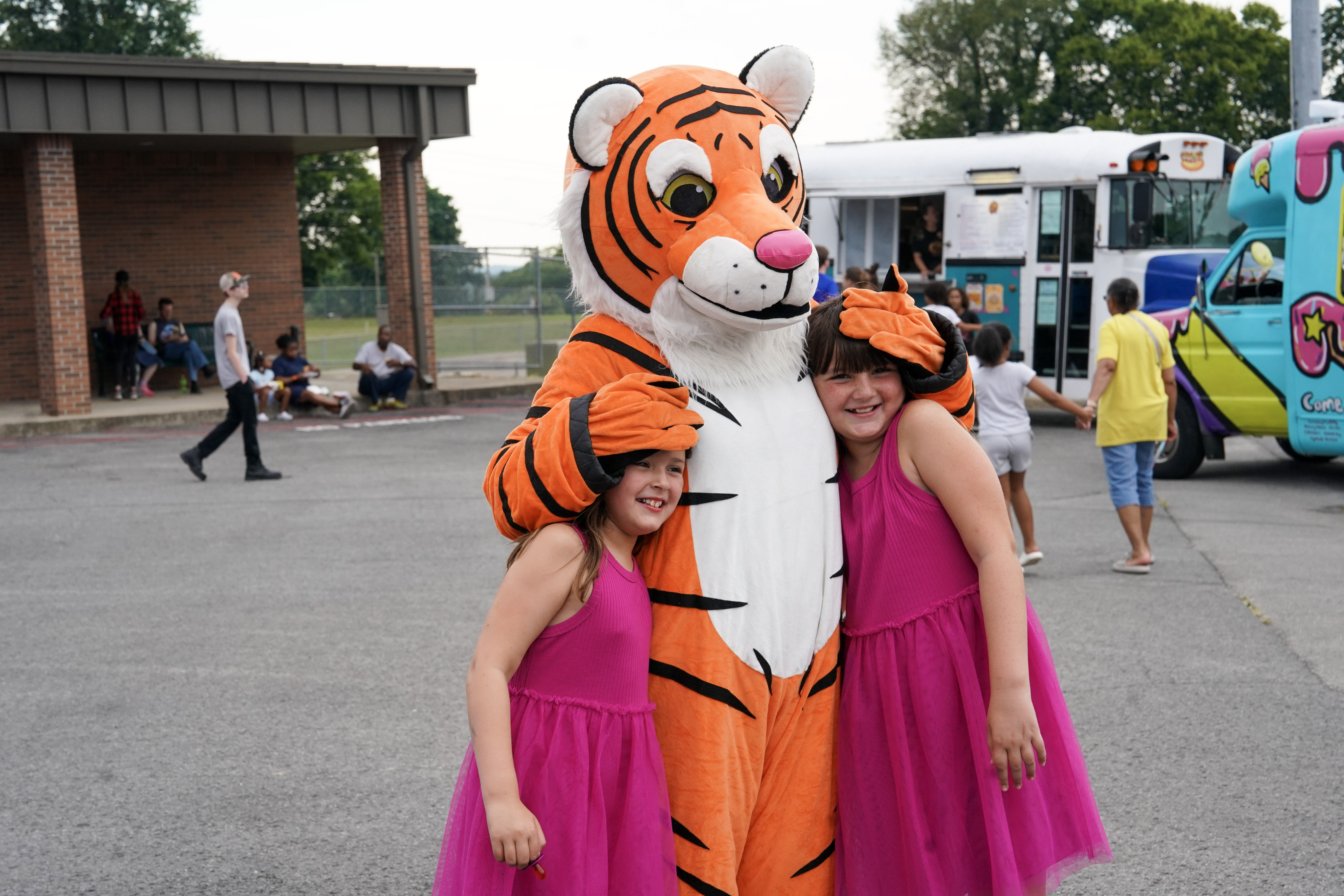 Two young girls in matching pink dresses smile while posing with a person in a friendly tiger costume at an outdoor event, with people, a bus, and an ice cream truck in the background.