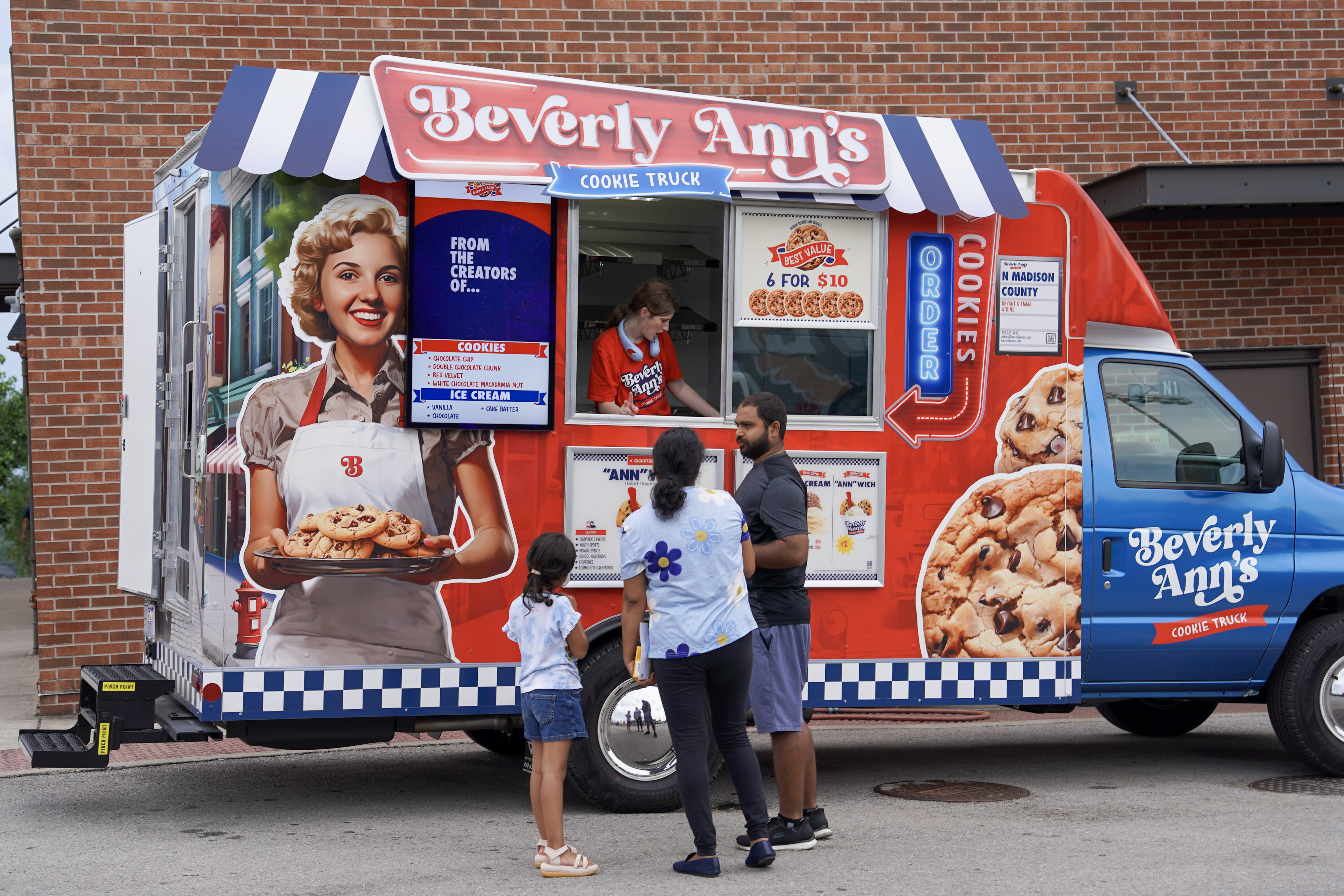 A family stands in front of a colorful “Beverly Ann’s Cookie Truck” parked on the street, ordering cookies from a worker inside. The truck is decorated with images of cookies and a smiling woman holding a tray.