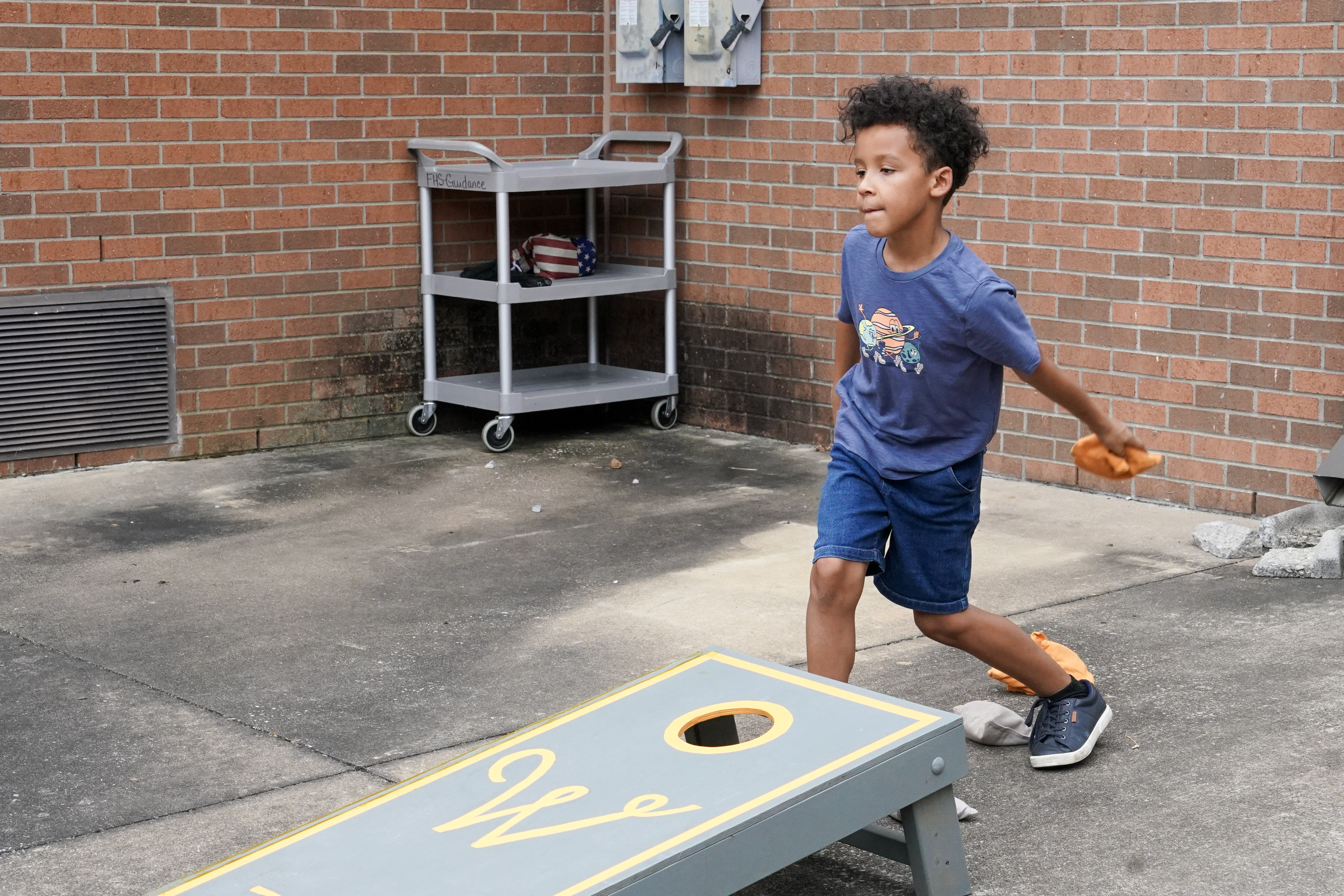 A young boy wearing a blue shirt and shorts tosses a bean bag while playing cornhole outside on a concrete surface near a brick wall.