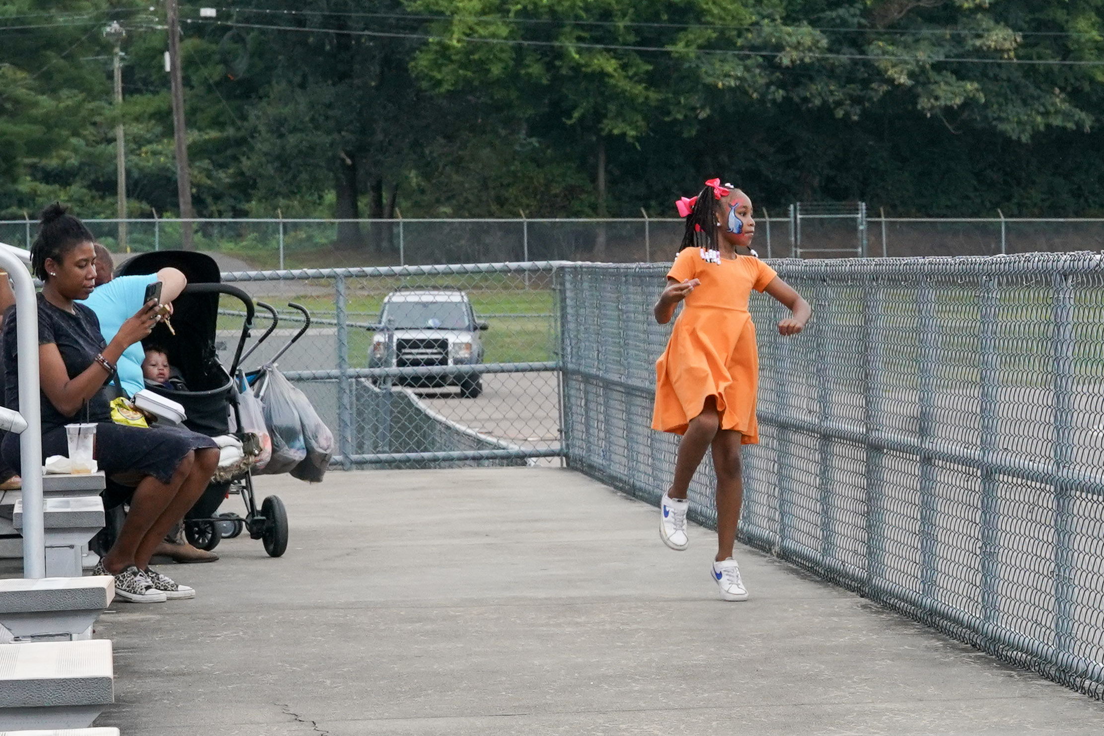 A young girl in an orange dress joyfully skips along a walkway near a chain-link fence, while a woman with a phone and a baby in a stroller sit on benches nearby.