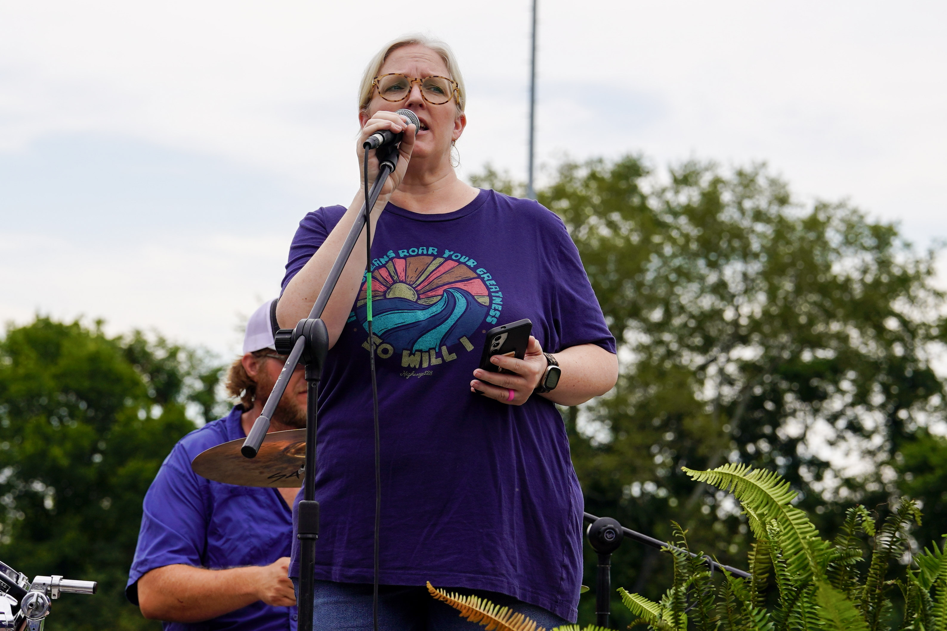 A person wearing glasses and a purple shirt sings into a microphone outdoors, holding a smartphone. Another person in a purple shirt and cap is partially visible behind them. Greenery and trees are in the background.