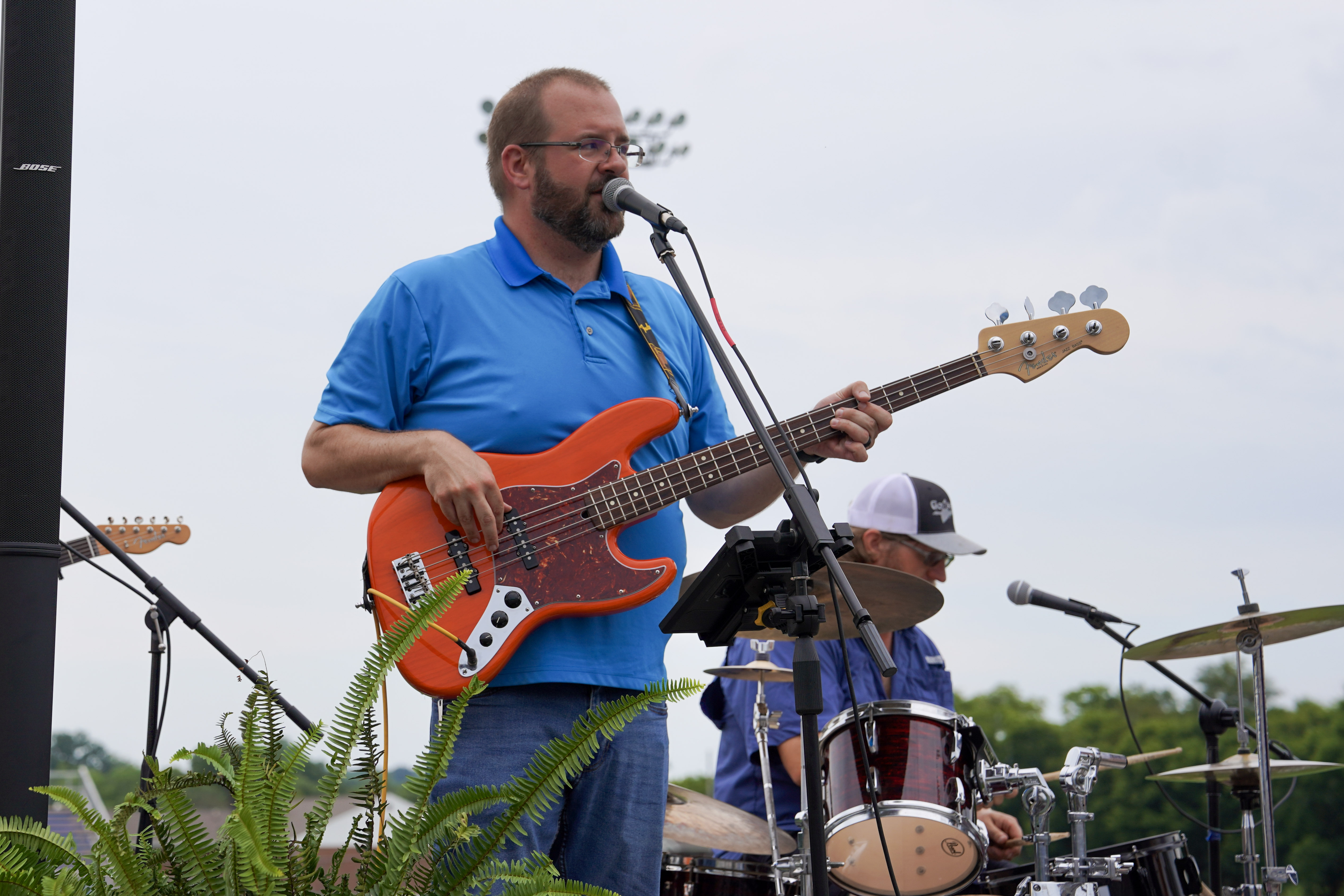 A man in a blue polo shirt plays an orange electric bass guitar and sings into a microphone on an outdoor stage, with a drummer and drum set visible in the background. A fern plant sits in front of the stage.