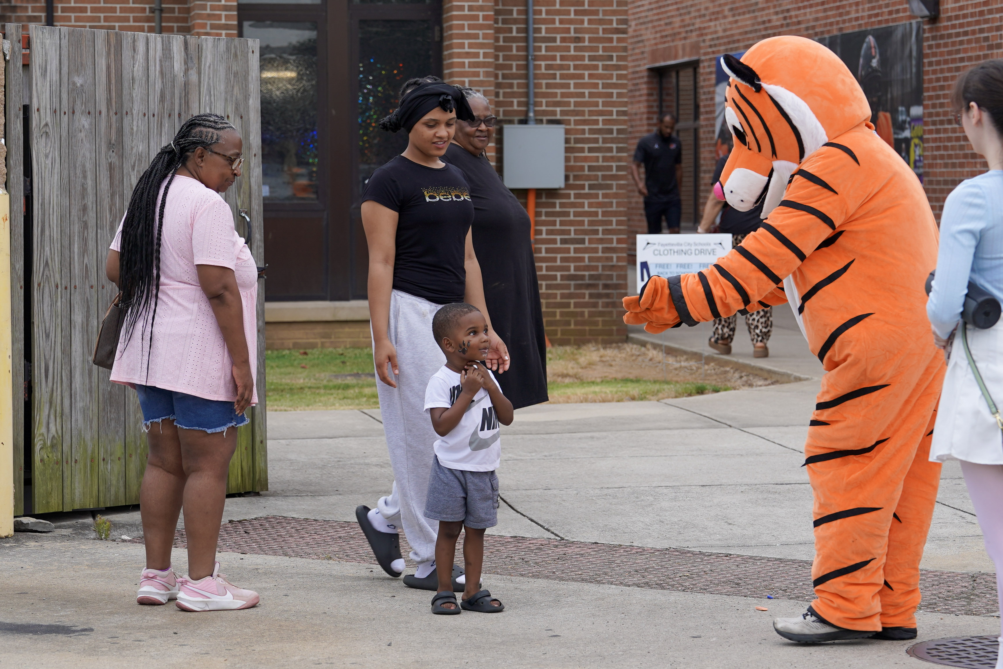 A person in a tiger costume greets a smiling young boy and a group of adults outside a brick building. The boy looks excited while the adults and another person stand nearby, watching the interaction.