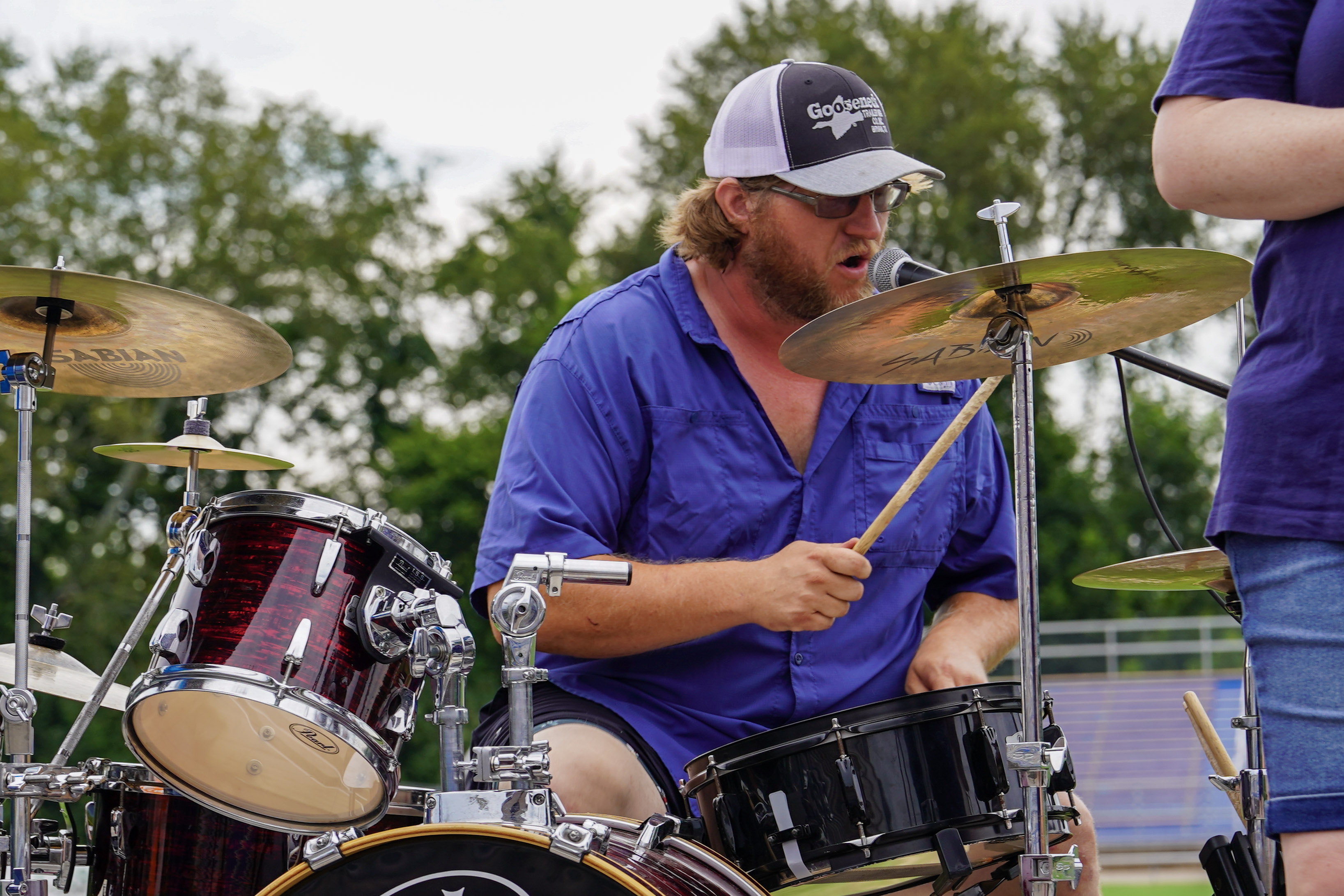 A man wearing a blue shirt and baseball cap plays drums outdoors, singing or speaking into a microphone. Trees and blurred bleachers are visible in the background.