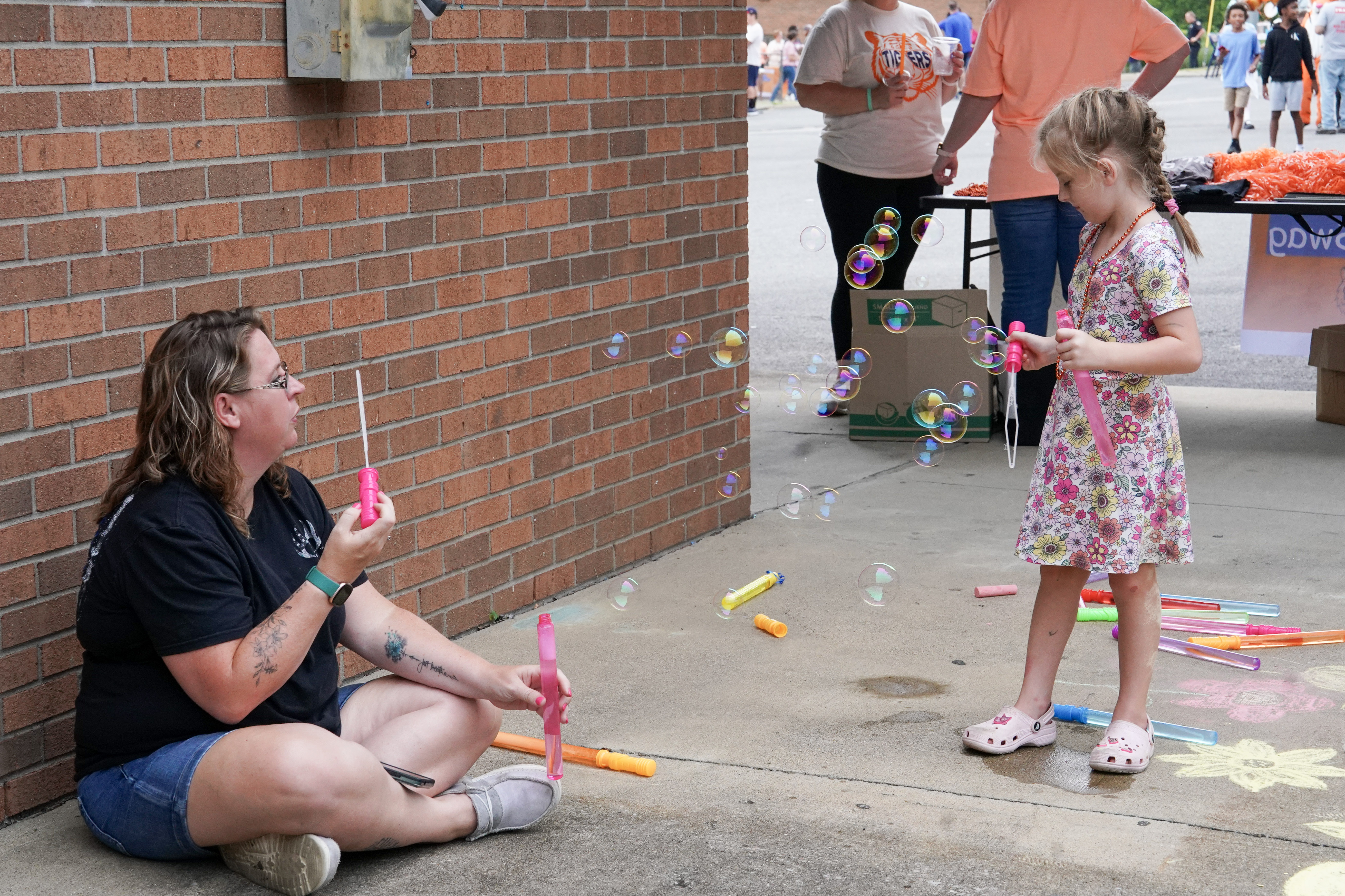 A woman sitting on the ground blows bubbles toward a young girl in a floral dress, who is holding a bubble wand. Colorful bubble wands are scattered nearby, and other people are visible in the background.