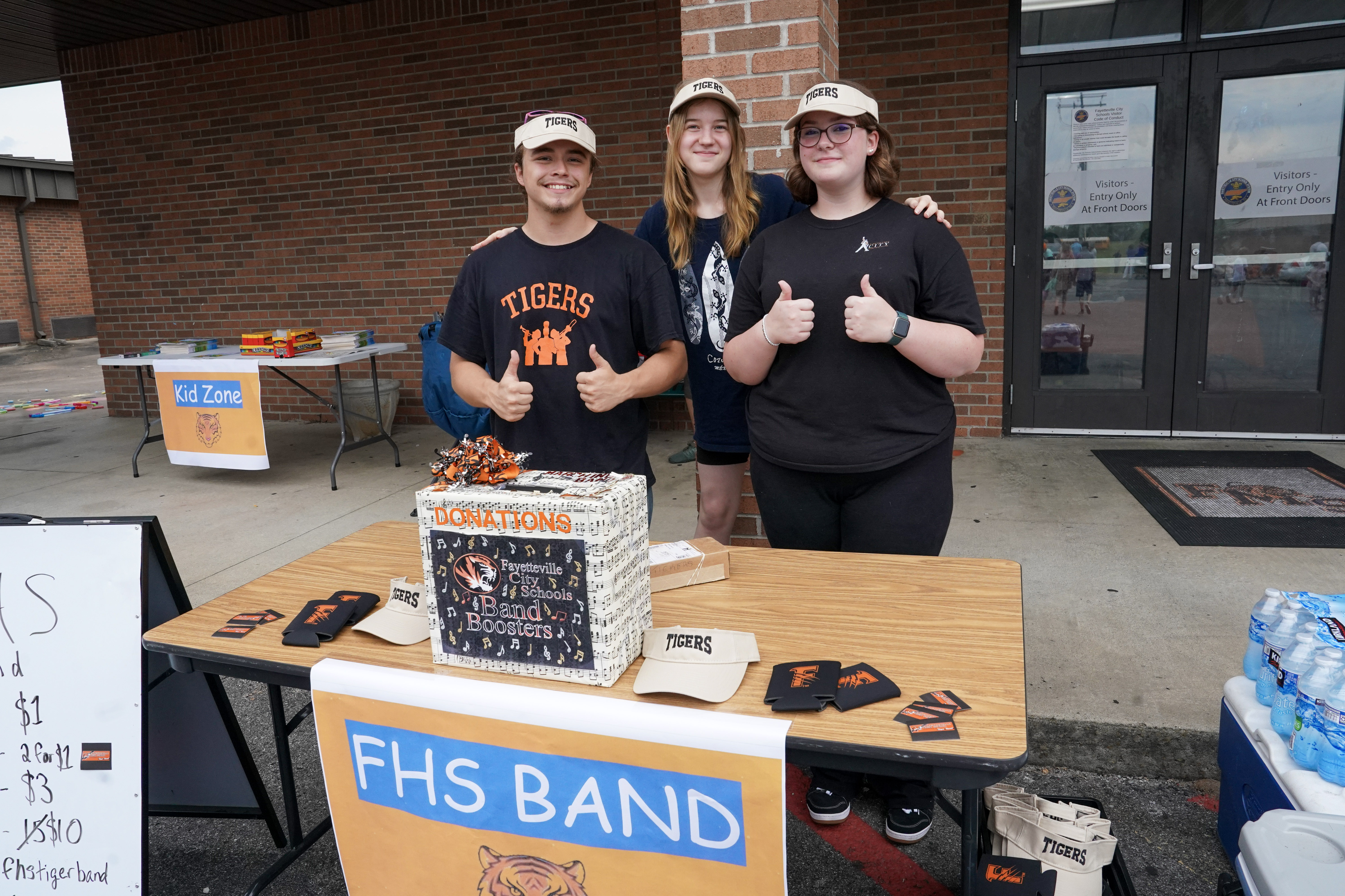 Three teens stand behind a table labeled FHS BAND with a decorated donation box, hats, and flyers; two give thumbs up. They wear matching hats and stand outside a brick building.