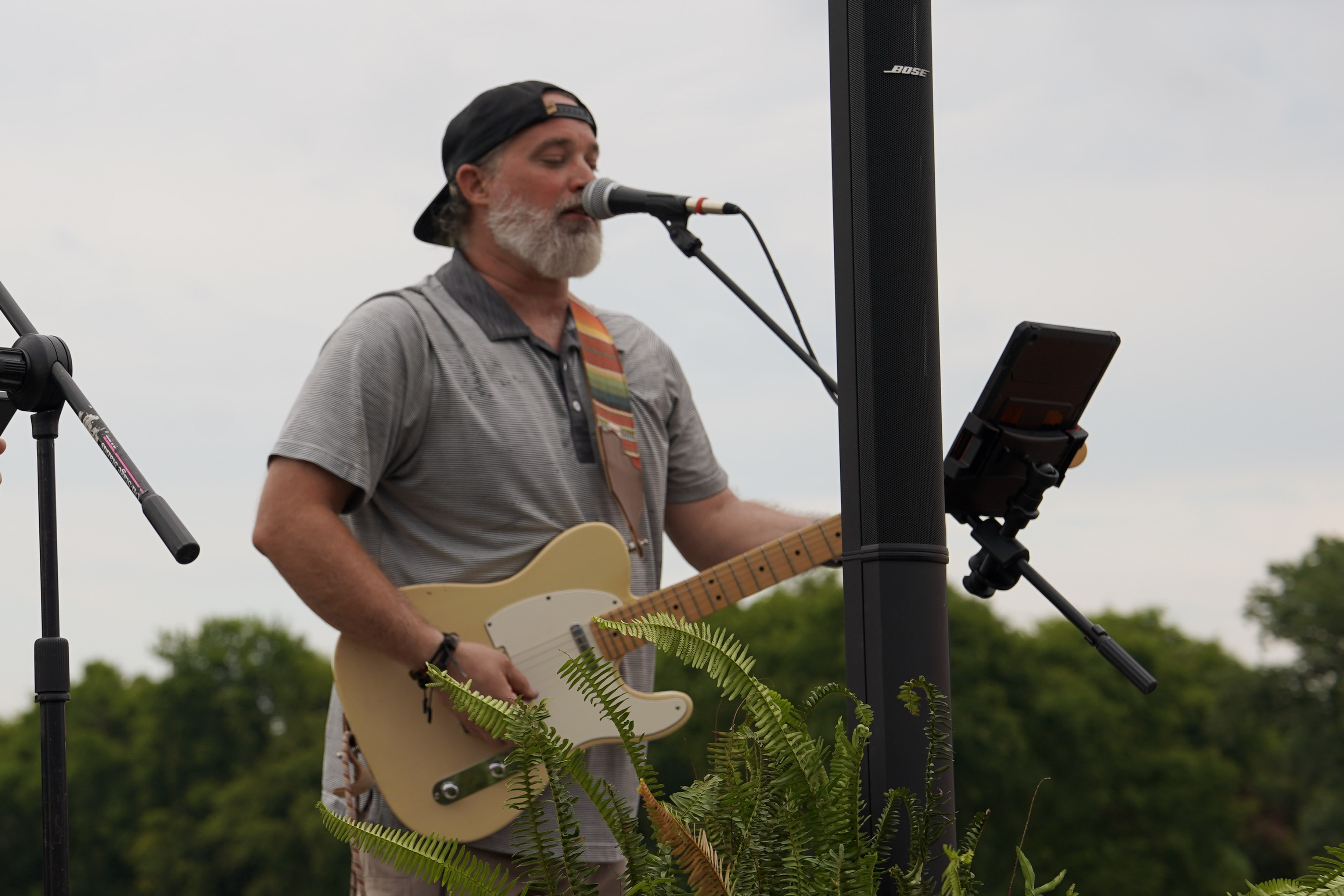 A man with a gray beard and backwards cap sings into a microphone and plays an electric guitar outdoors, standing behind a plant with trees in the background.