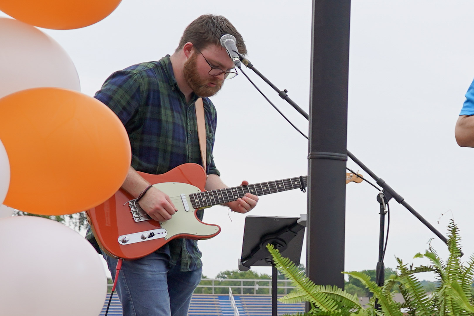 A man in a plaid shirt plays an electric guitar on an outdoor stage. Orange and white balloons are on the left, and a microphone, music stand, and green ferns are in the foreground. The sky is overcast.