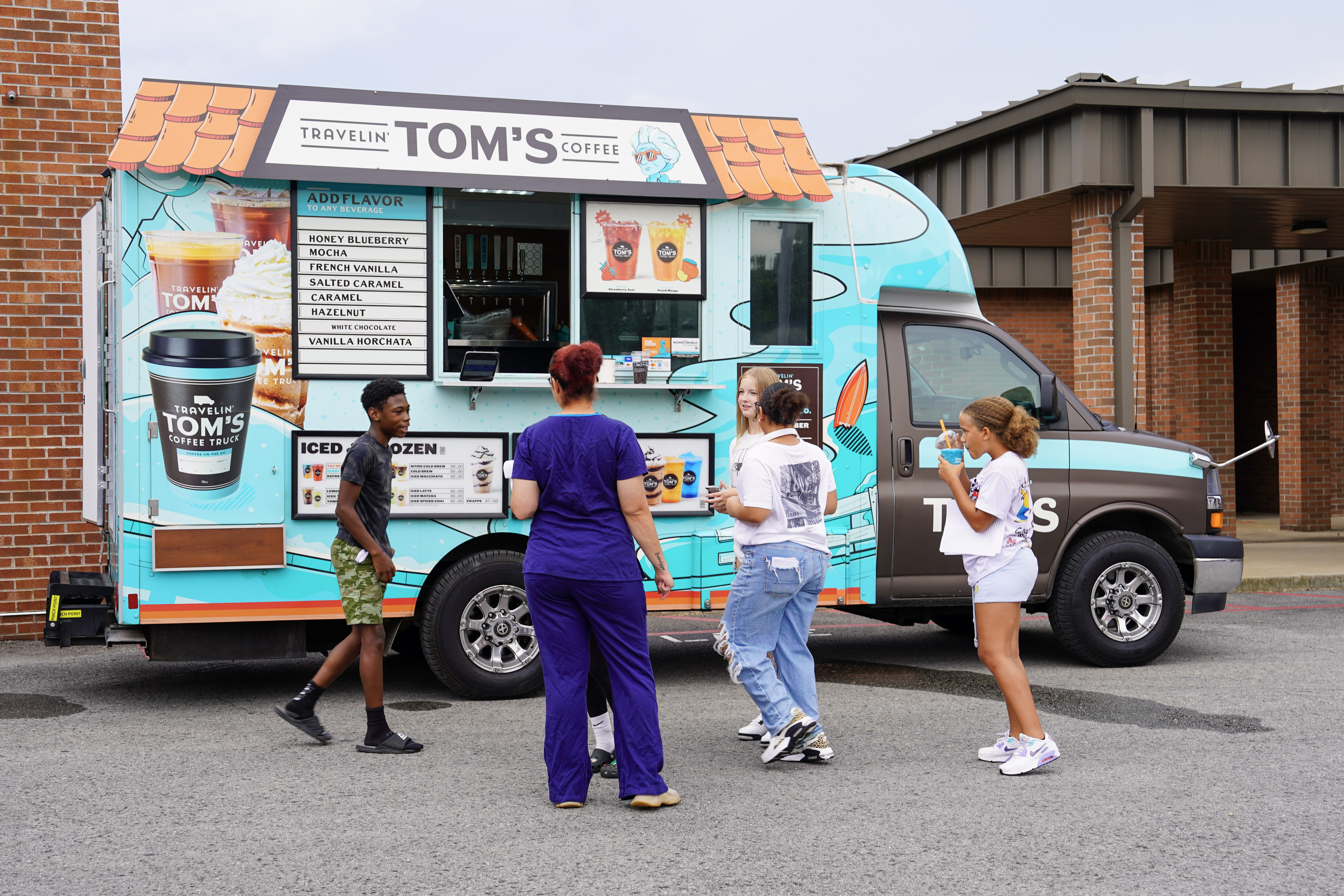 Four people stand near a colorful coffee truck labeled Travelin Toms Coffee, parked by a brick building, as they look at the menu and prepare to order drinks.