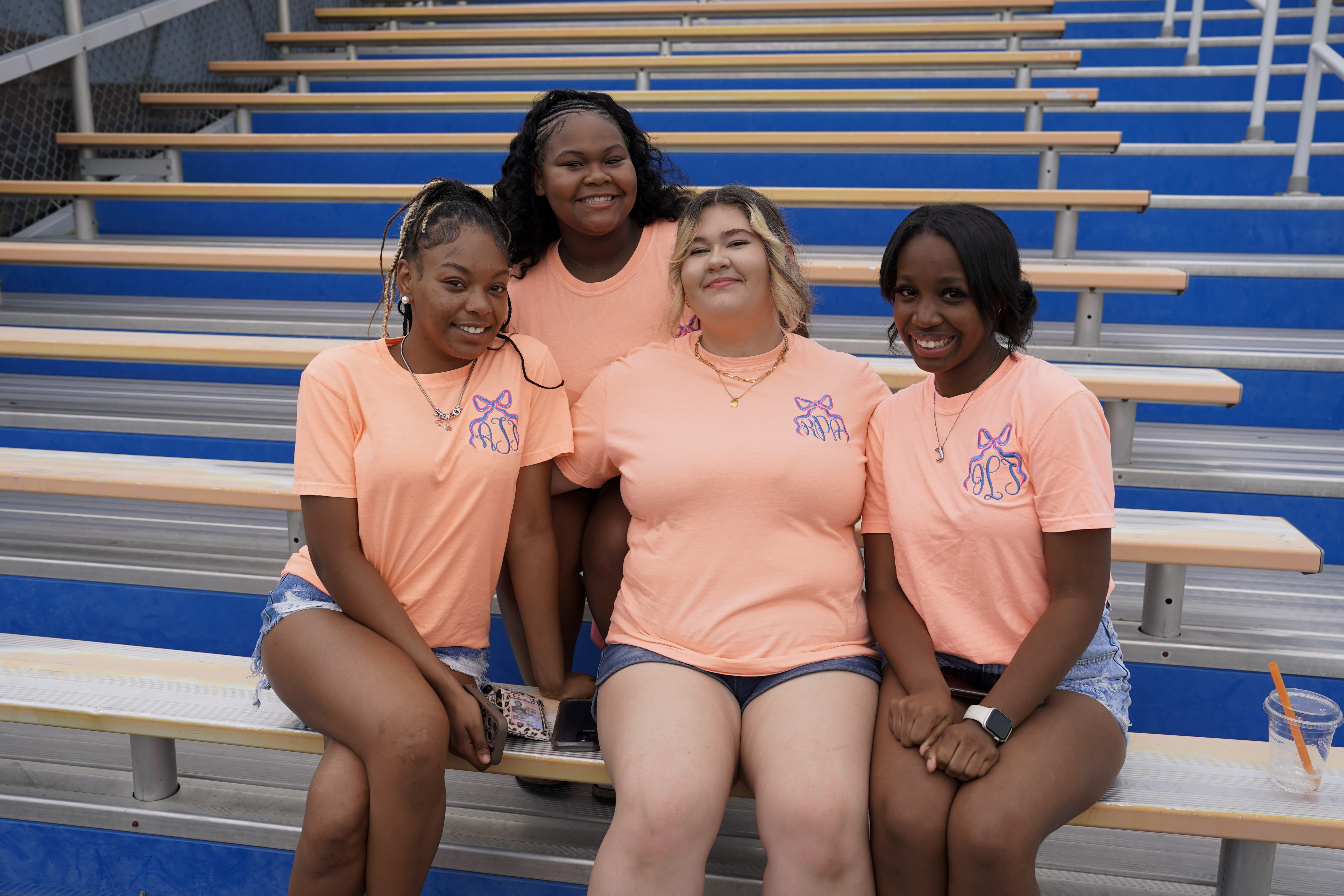Four young women sit together on bleachers, smiling at the camera. They are wearing matching peach T-shirts and denim shorts. The bleachers are blue and silver, and an empty drink cup is placed nearby.