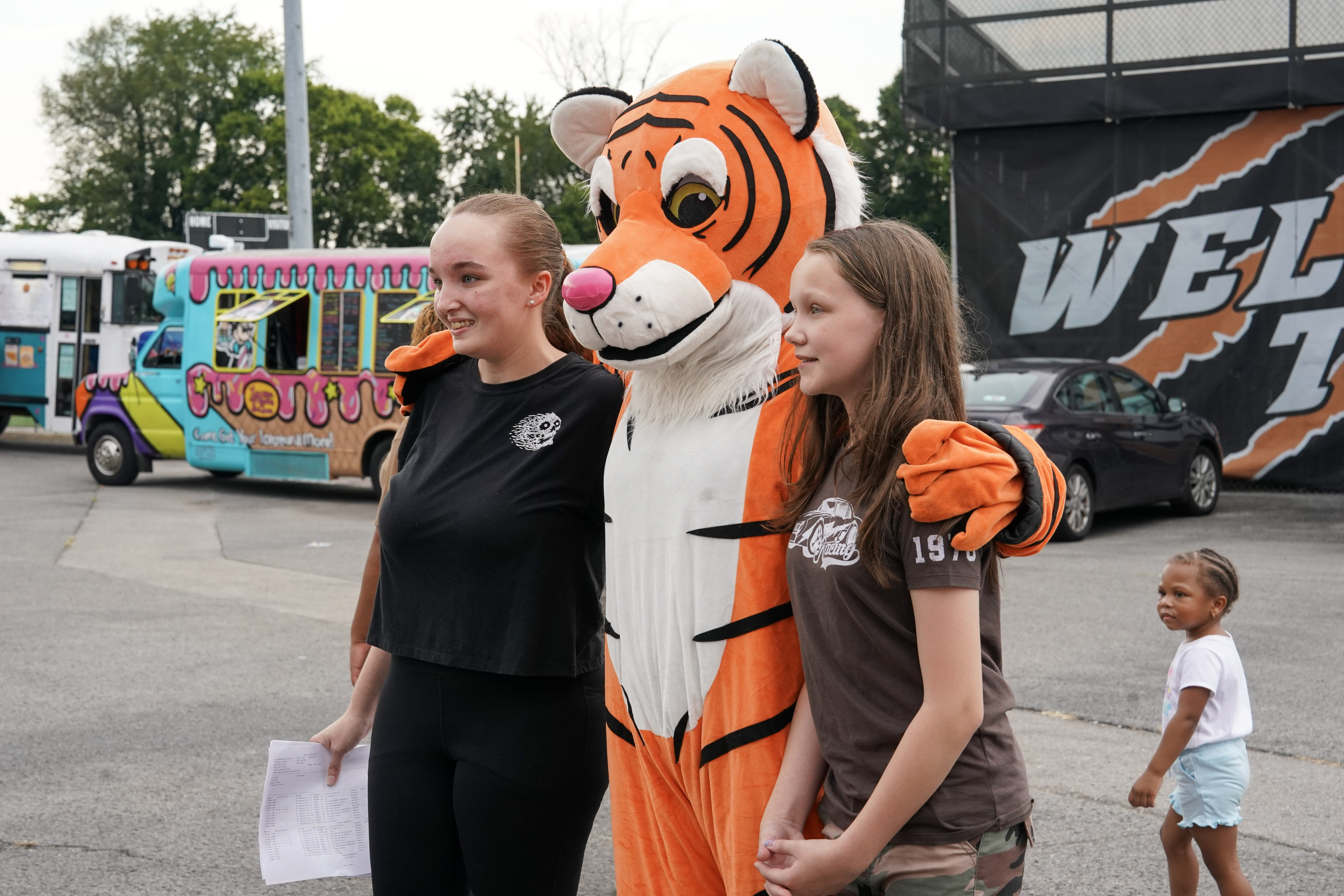 Two girls pose for a photo with a person in a tiger mascot costume outside at an event. An ice cream truck and parked cars are visible in the background, with another young child walking nearby.