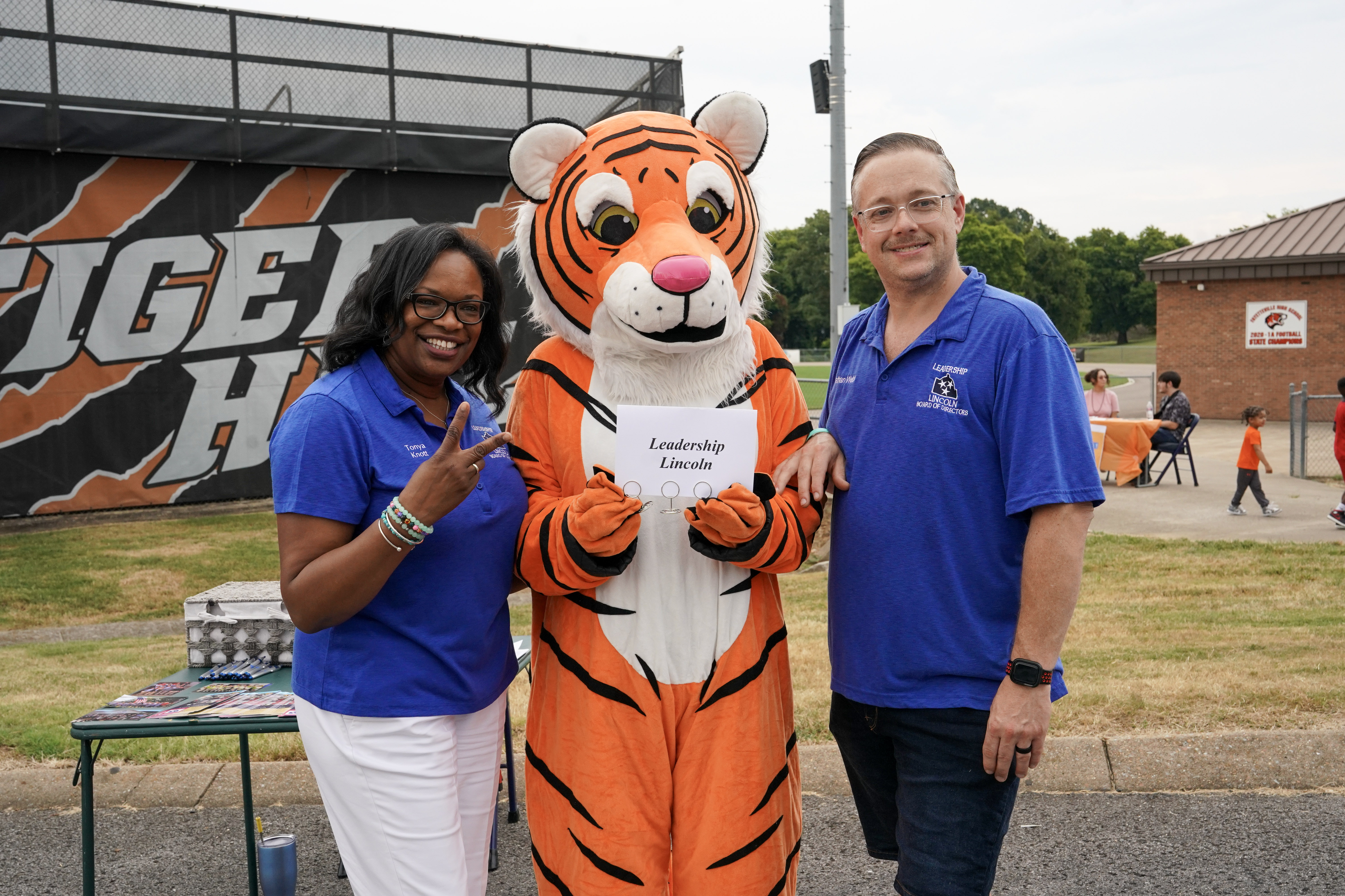 Two adults in blue shirts pose with a person in a tiger mascot costume holding a Leadership Lincoln sign. They are outdoors near a school with a Tiger banner in the background.