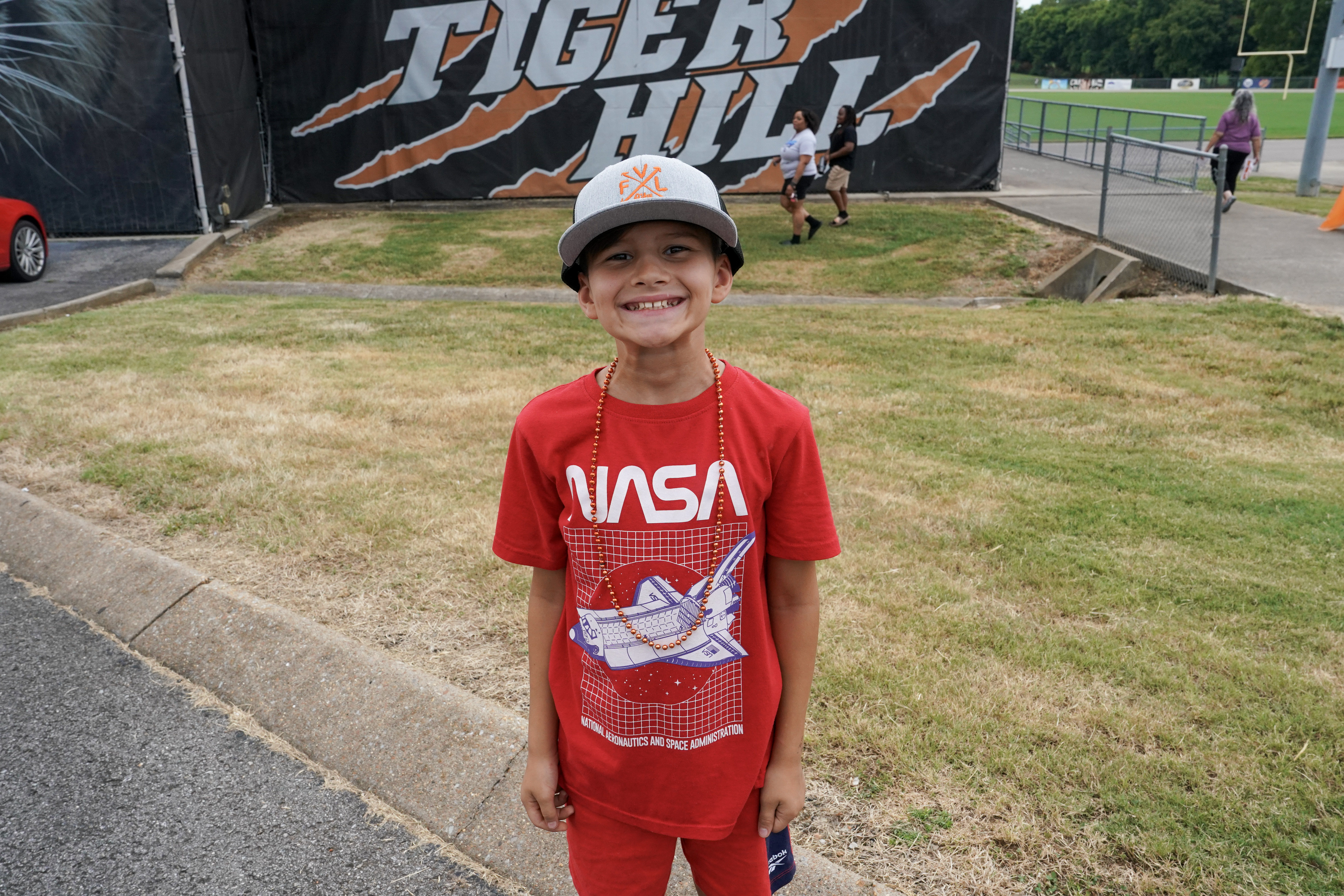 A smiling boy wearing a NASA shirt, red shorts, and a gray cap stands on grass in front of a Tiger Hill sign at an outdoor sports facility.