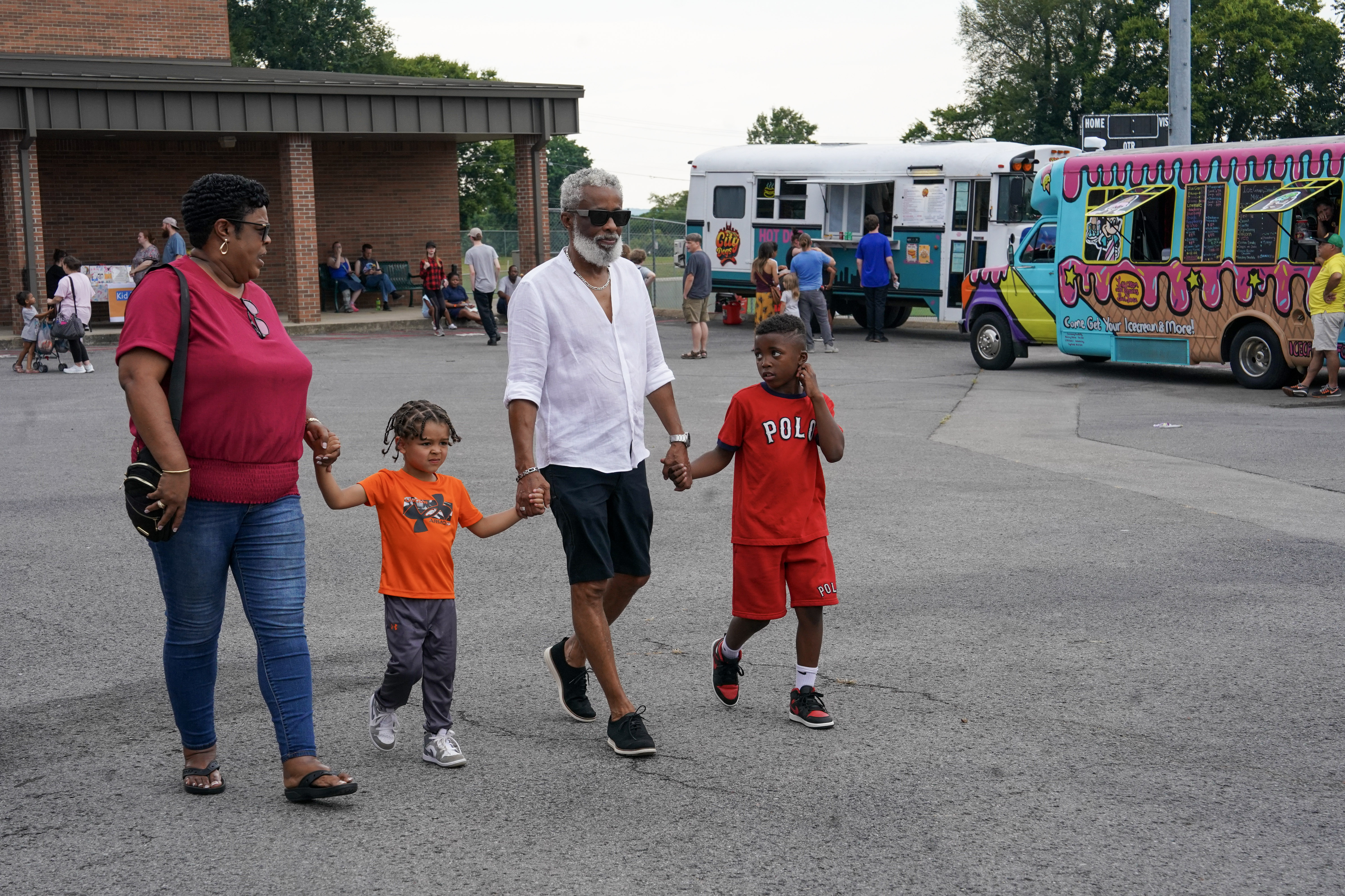 A family of four walks in a parking lot near an ice cream truck, holding hands. Other people stand nearby, and a colorful food truck is in the background on a sunny day.