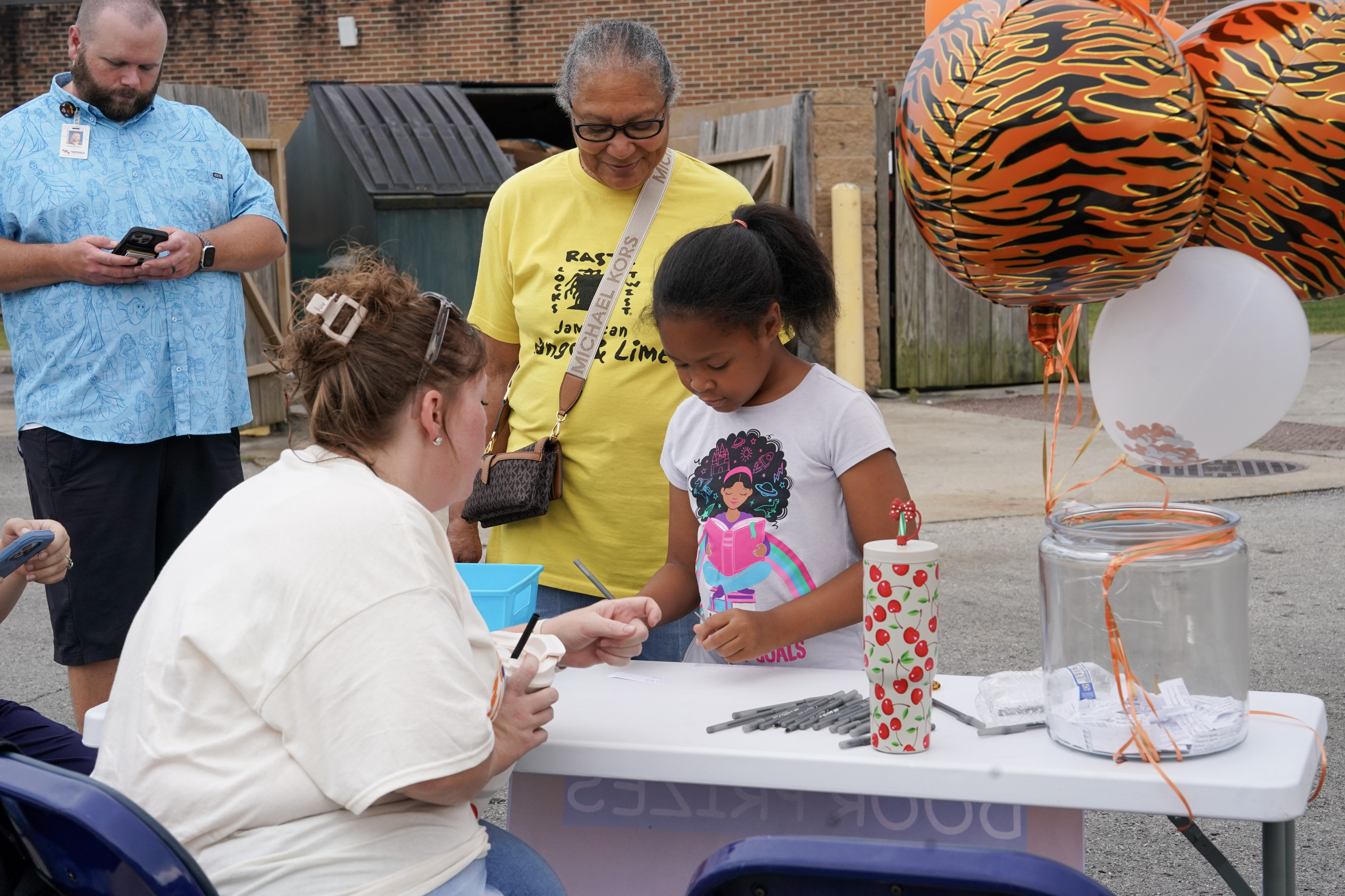 A woman sits at a table with pens and a raffle box, helping a young girl fill out a ticket. An older woman and another man stand nearby. Orange and tiger-striped balloons decorate the table.