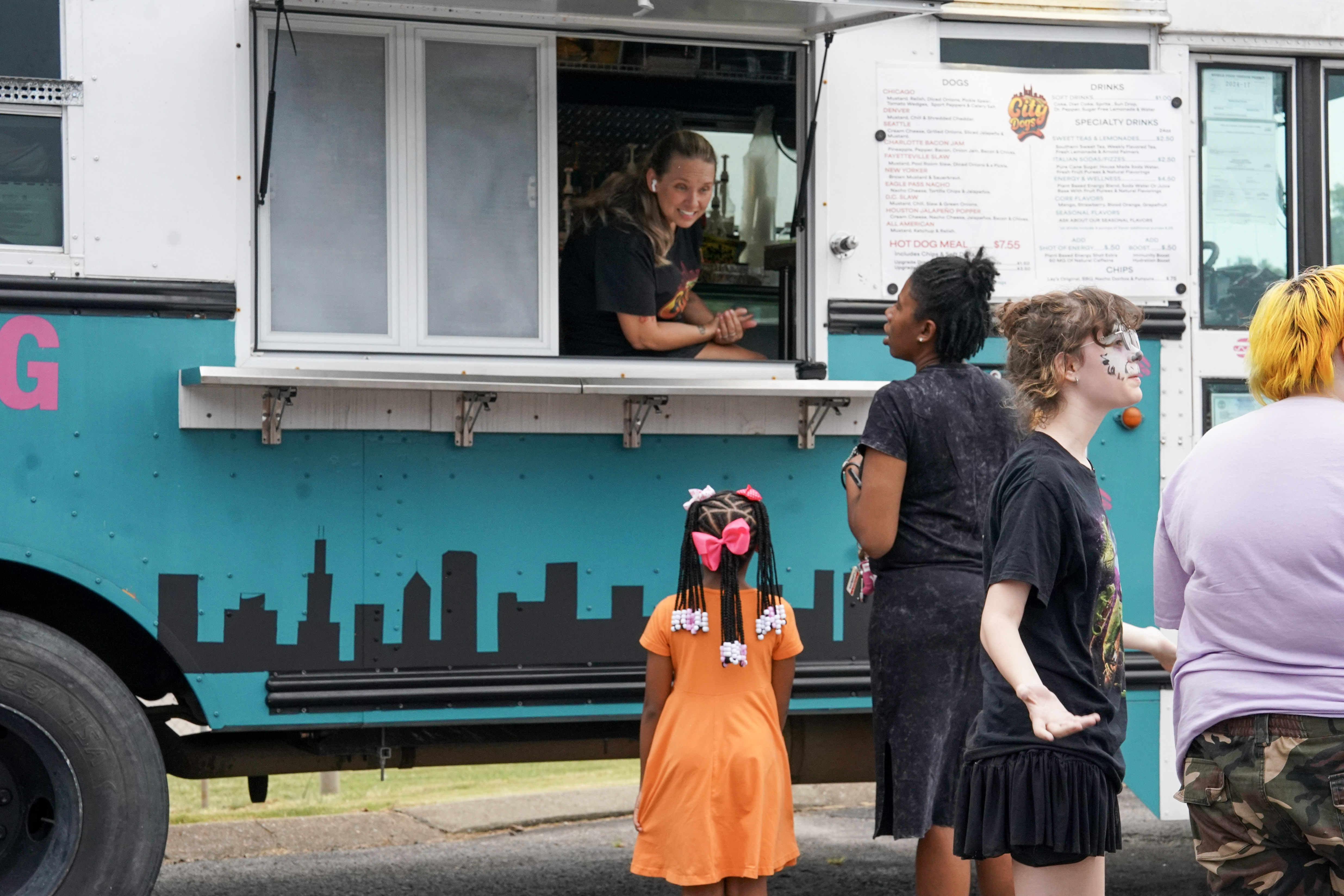 A woman smiles while serving customers from a food truck window. Several people, including children in colorful outfits and face paint, stand in line outdoors near the truck decorated with a city skyline.