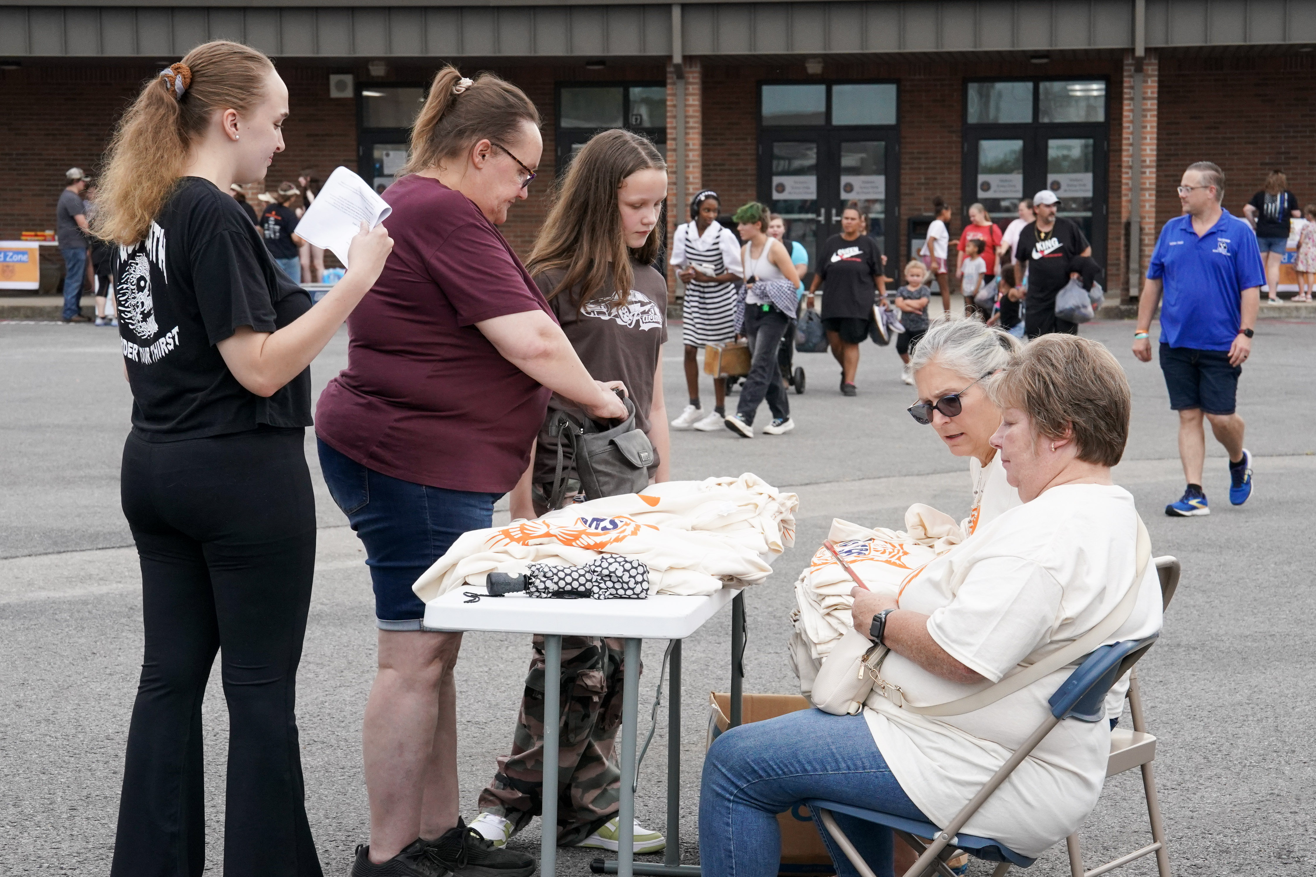 Several people gather around a table with folded t-shirts outside a building; two women sit while others stand and look at the shirts. More people are visible in the background.