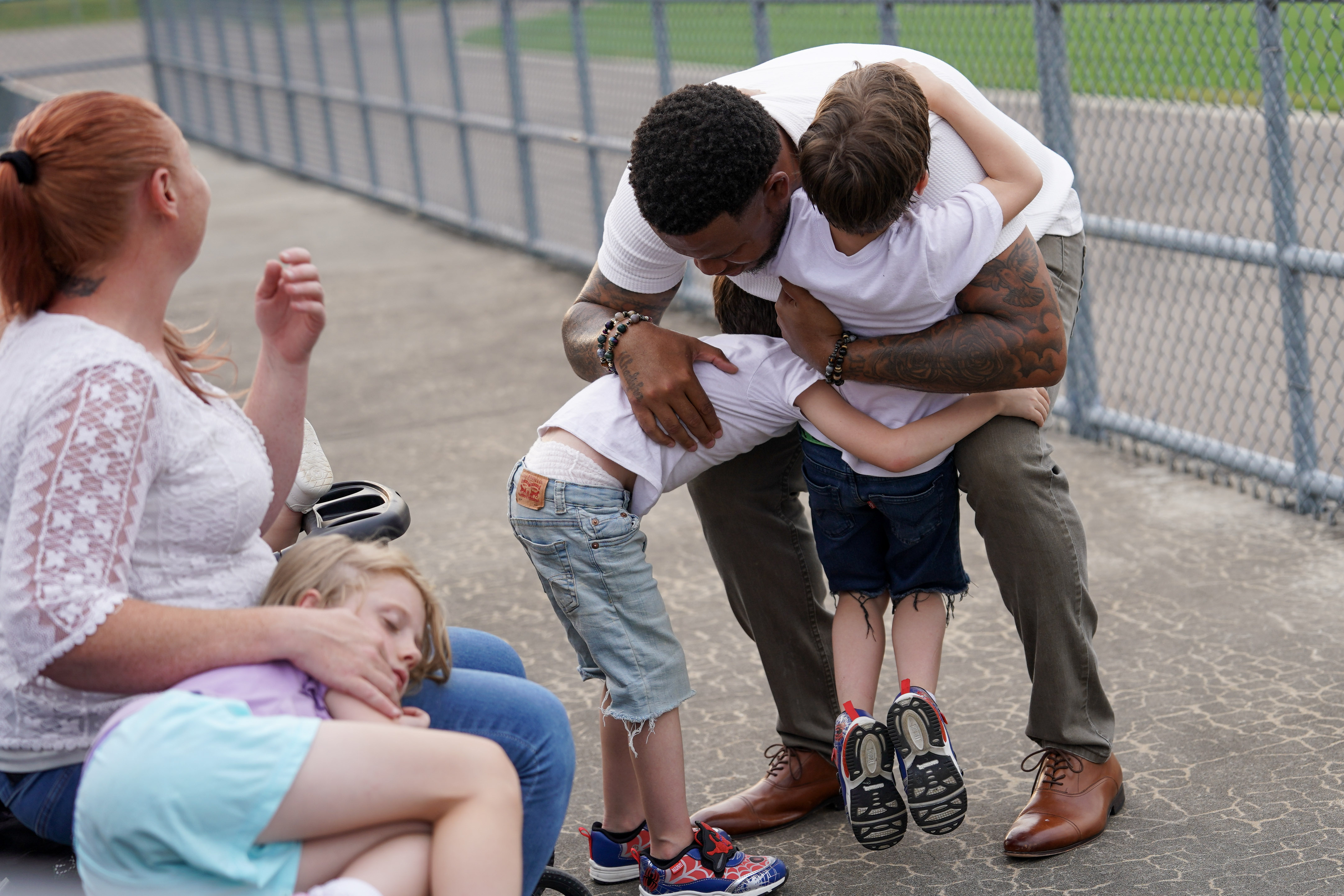 A man hugs two young boys tightly while a woman and a girl sit nearby on a bench. The setting is outdoors near a chain-link fence on a paved surface. The mood is warm and affectionate.