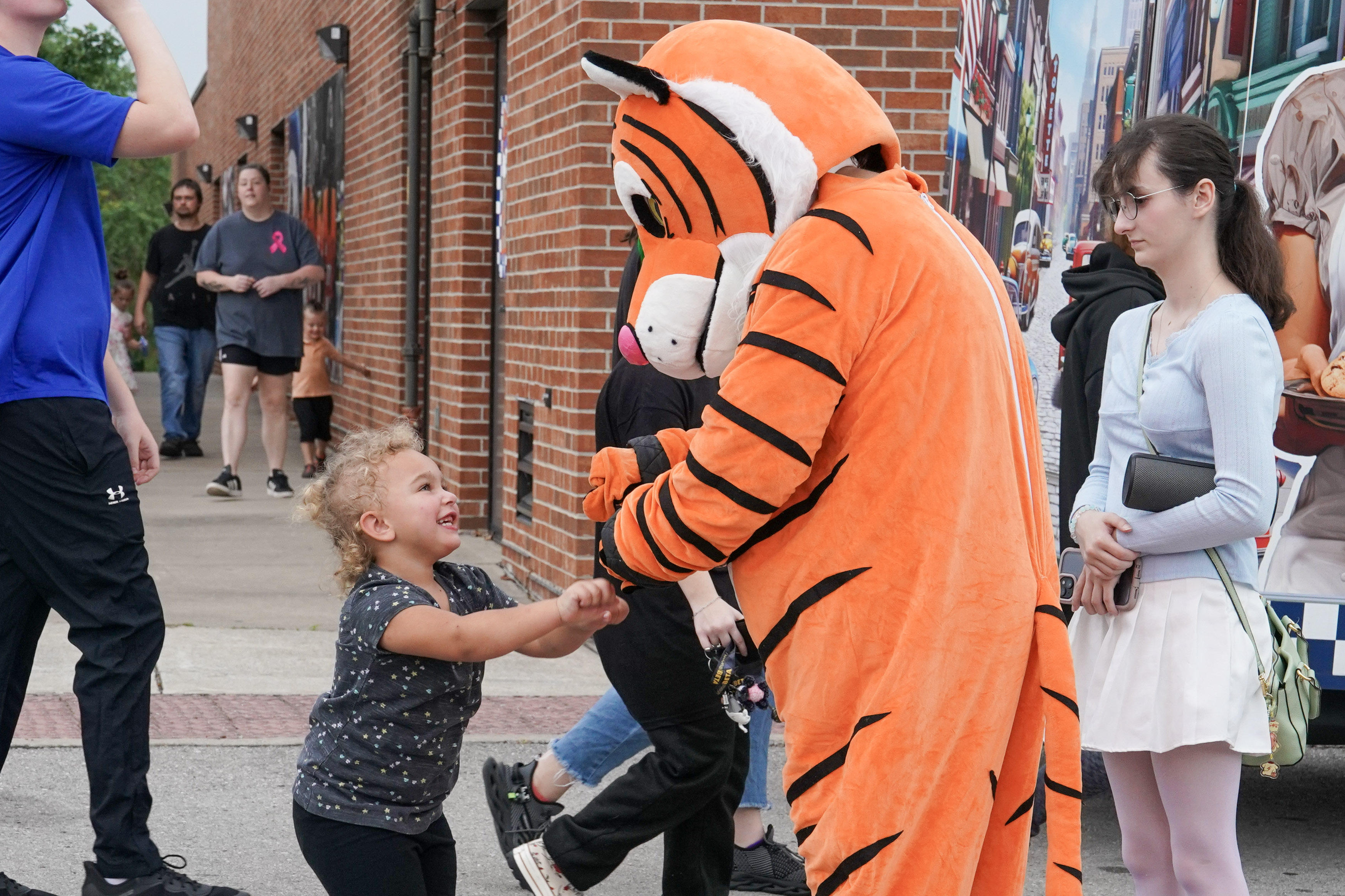 A young child smiles and holds hands with a person in a tiger costume outdoors, while other people walk by on the sidewalk near a brick building.