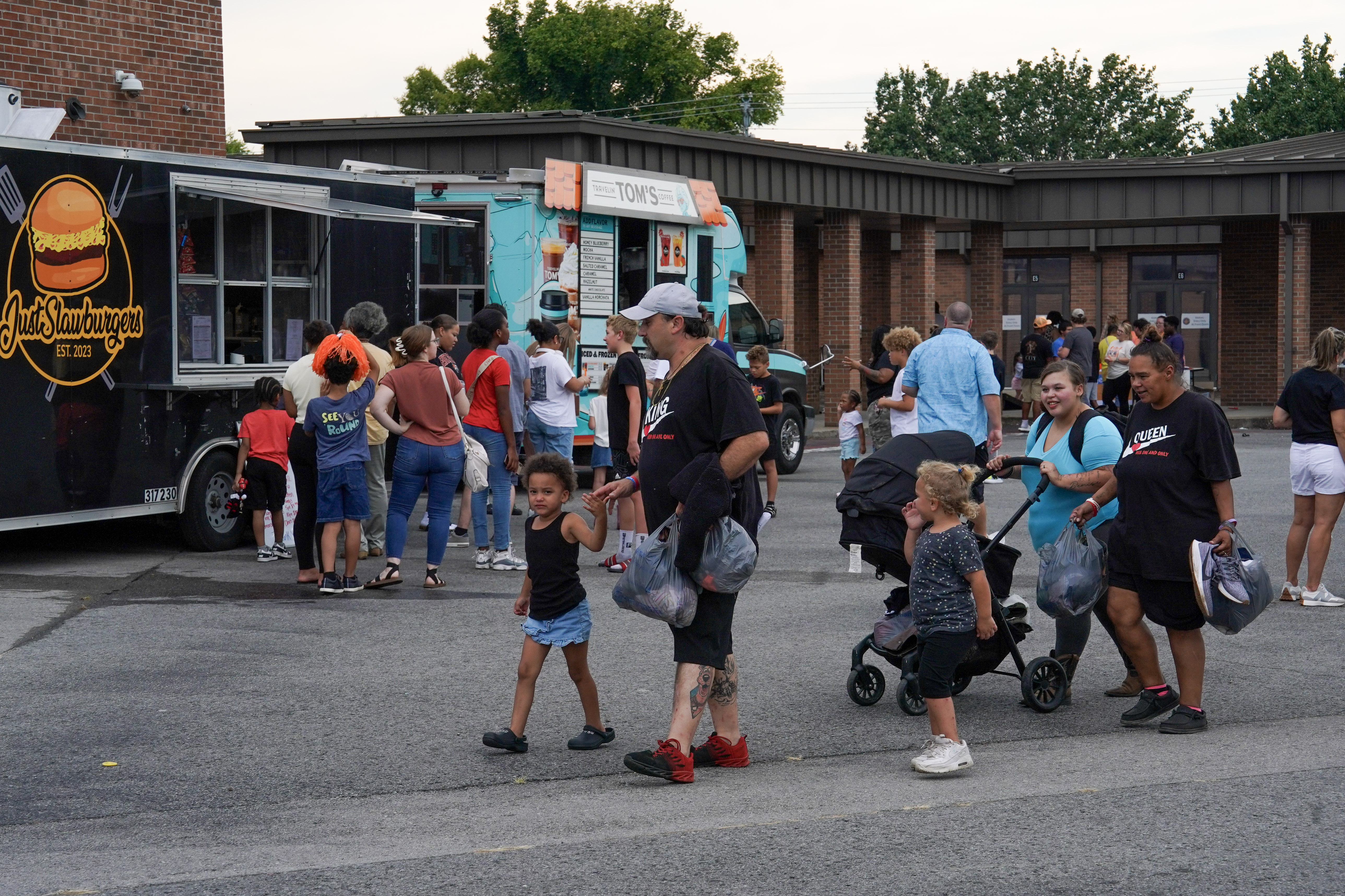 People gather around food trucks in a parking lot; some wait in line while others walk away carrying bags of food. Children and adults are present, and a few people push strollers. Trees and buildings are in the background.