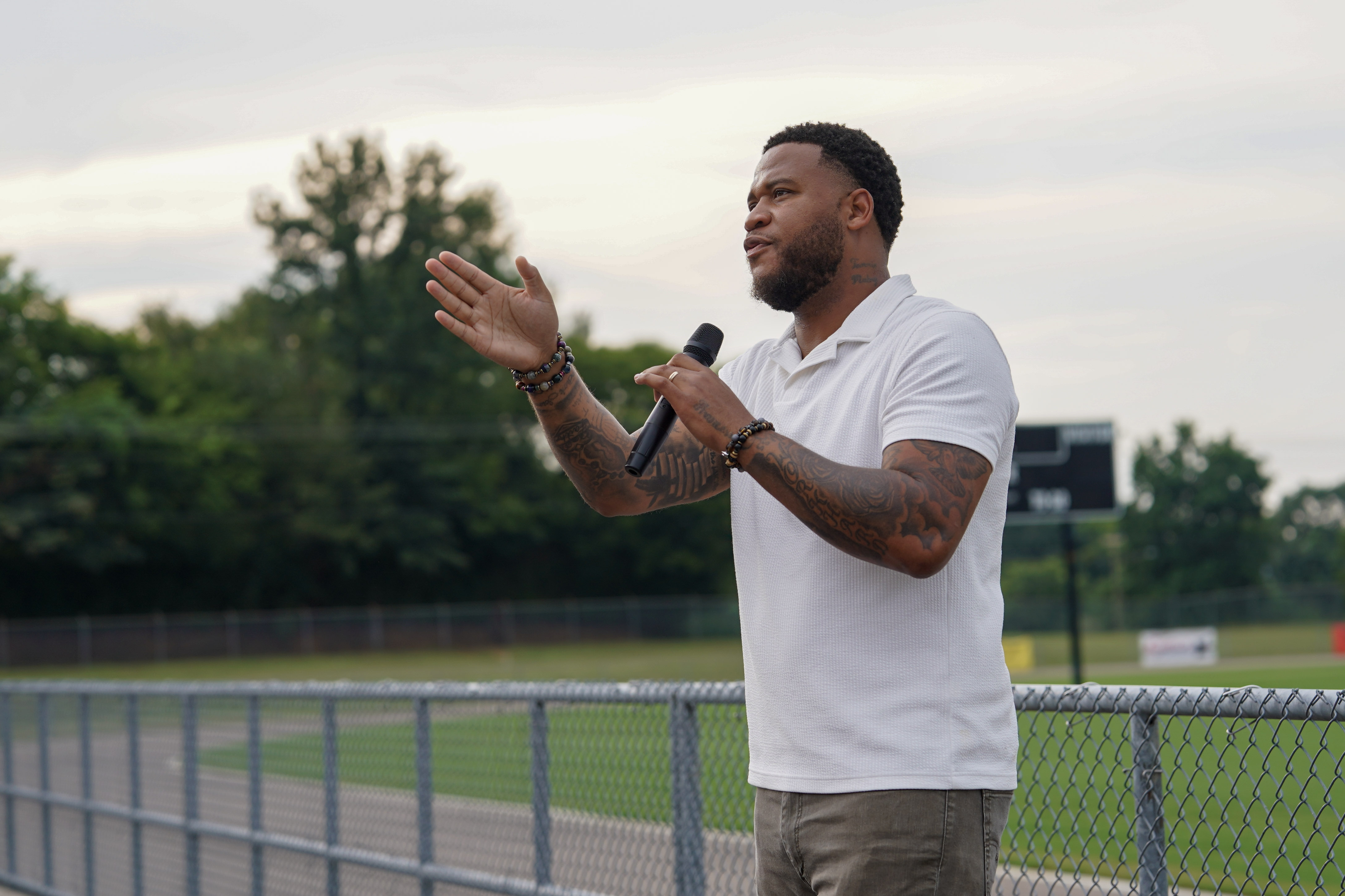 A man with tattoos wearing a white polo shirt speaks into a microphone while gesturing with his hand, standing outdoors by a chain-link fence on a sports field. Trees and a scoreboard are visible in the background.