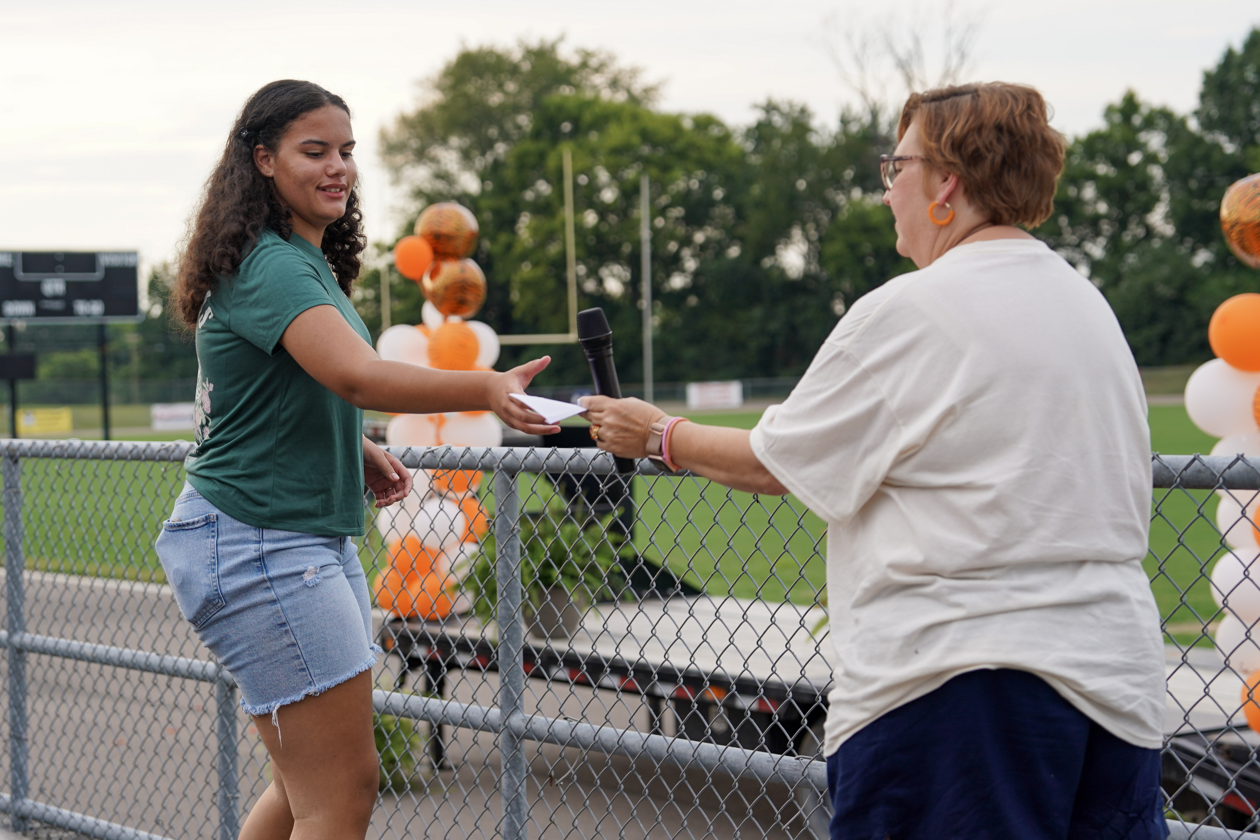 A young woman in a green shirt and denim shorts receives a microphone from an older woman near a sports field decorated with orange and white balloons.