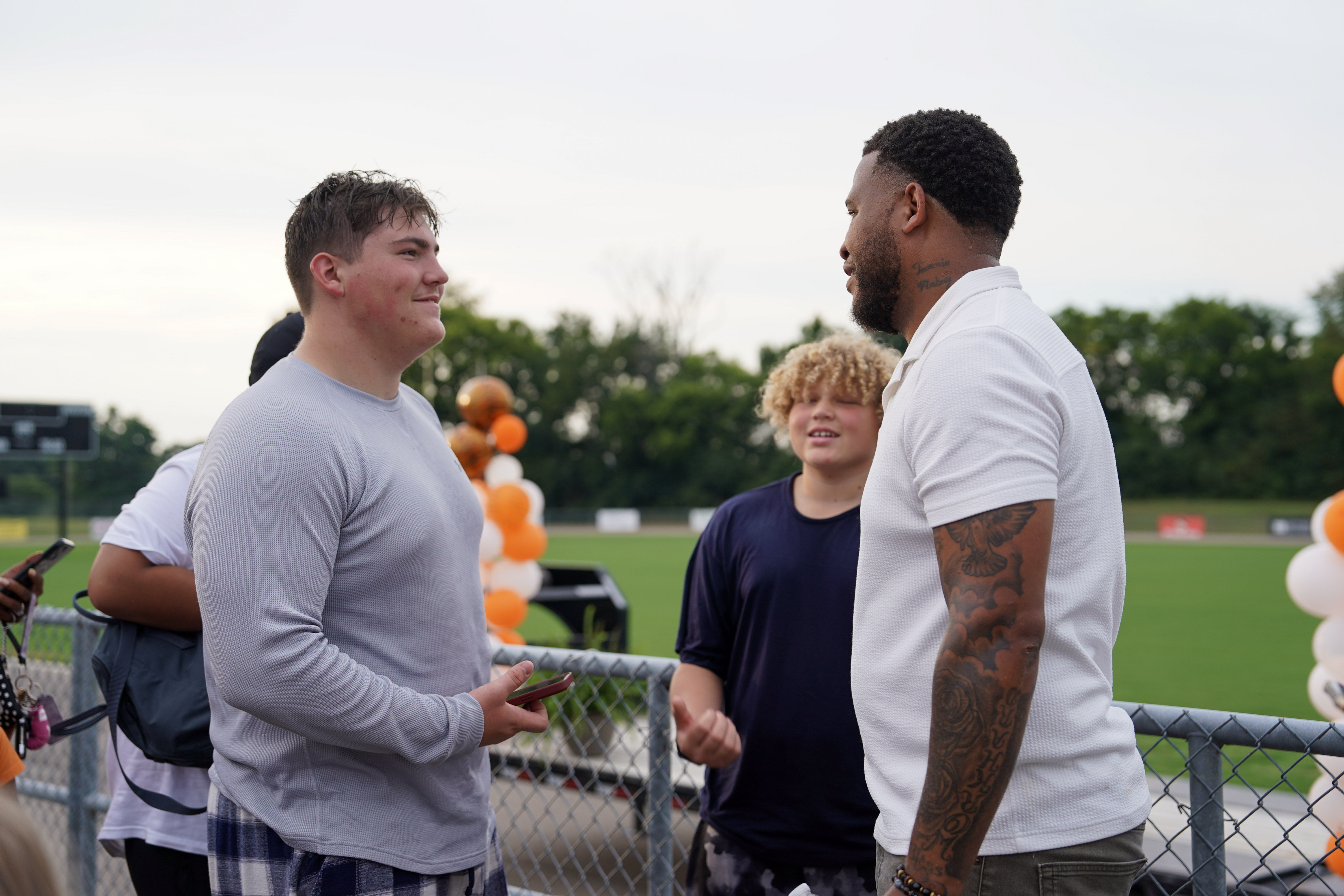 A man with tattoos on his arm talks with two teenage boys by a football field. One boy holds a phone and smiles, while another stands in the background. Orange and white balloons decorate the fence nearby.