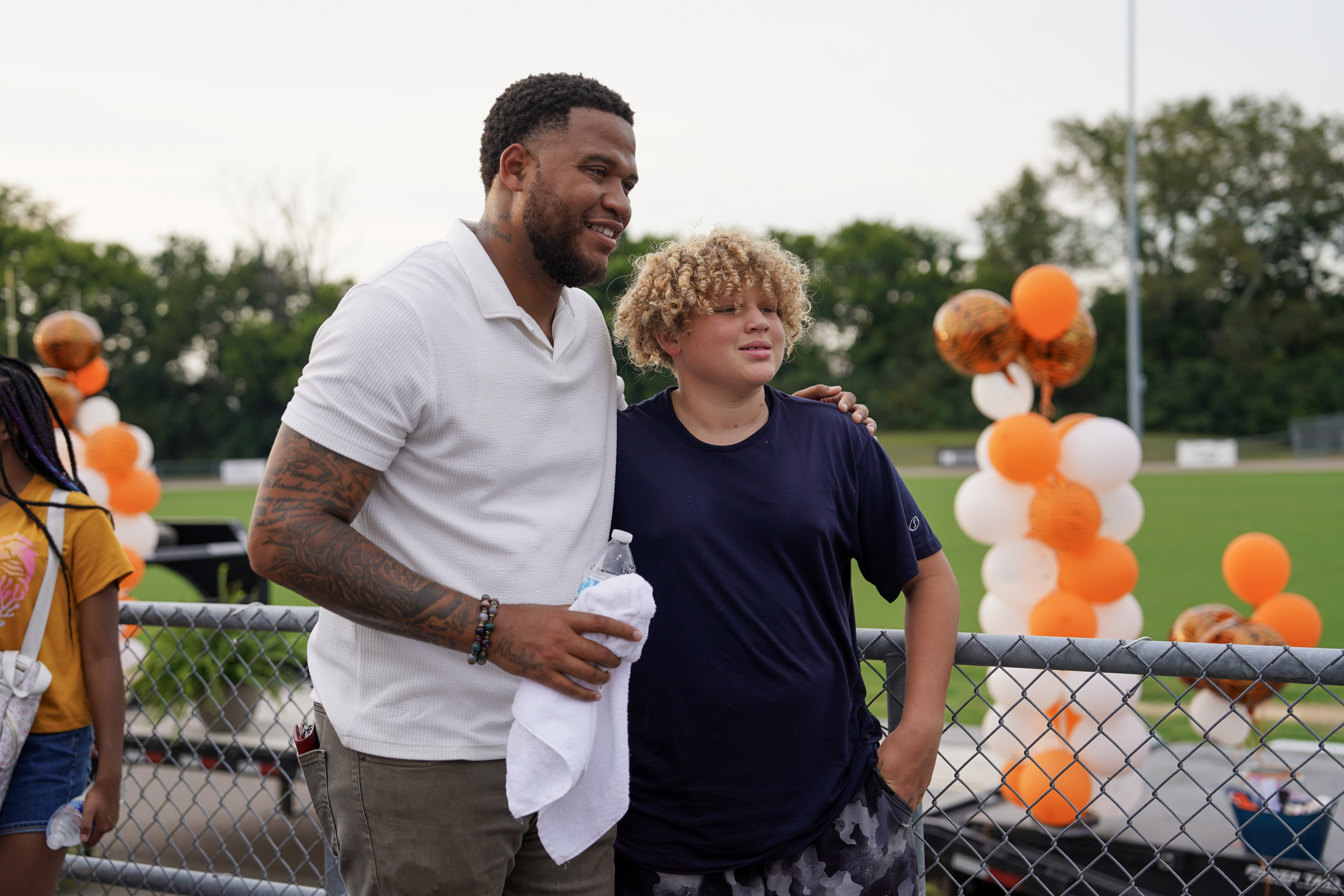 A man in a white shirt stands with his arm around a smiling boy wearing a navy shirt. They are outdoors near a fence, with orange and white balloons and greenery in the background.