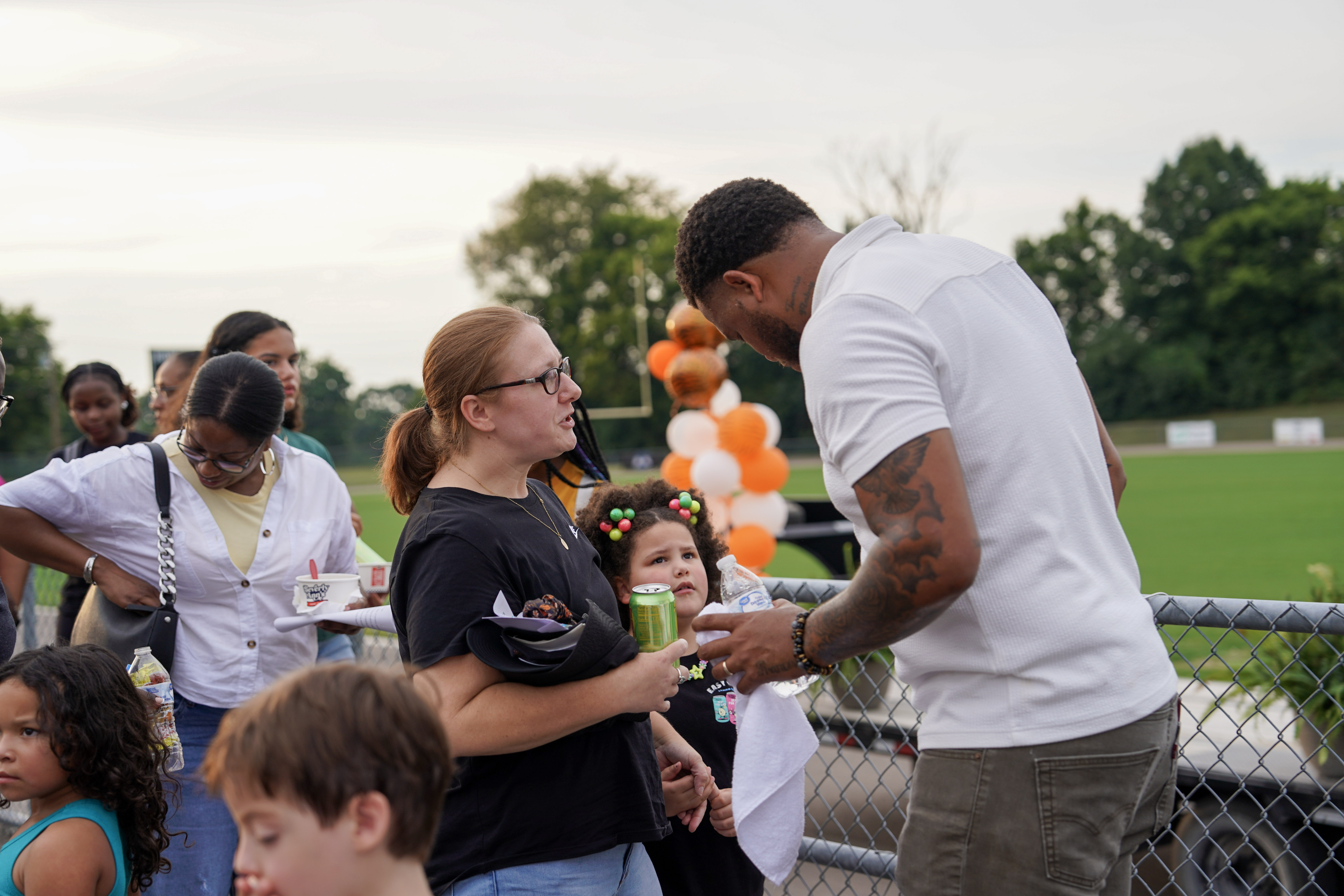A group of adults and children gather outdoors near a chain-link fence, with balloons and a green field in the background. One woman holds a drink and a small child, while a man in a white shirt looks down.