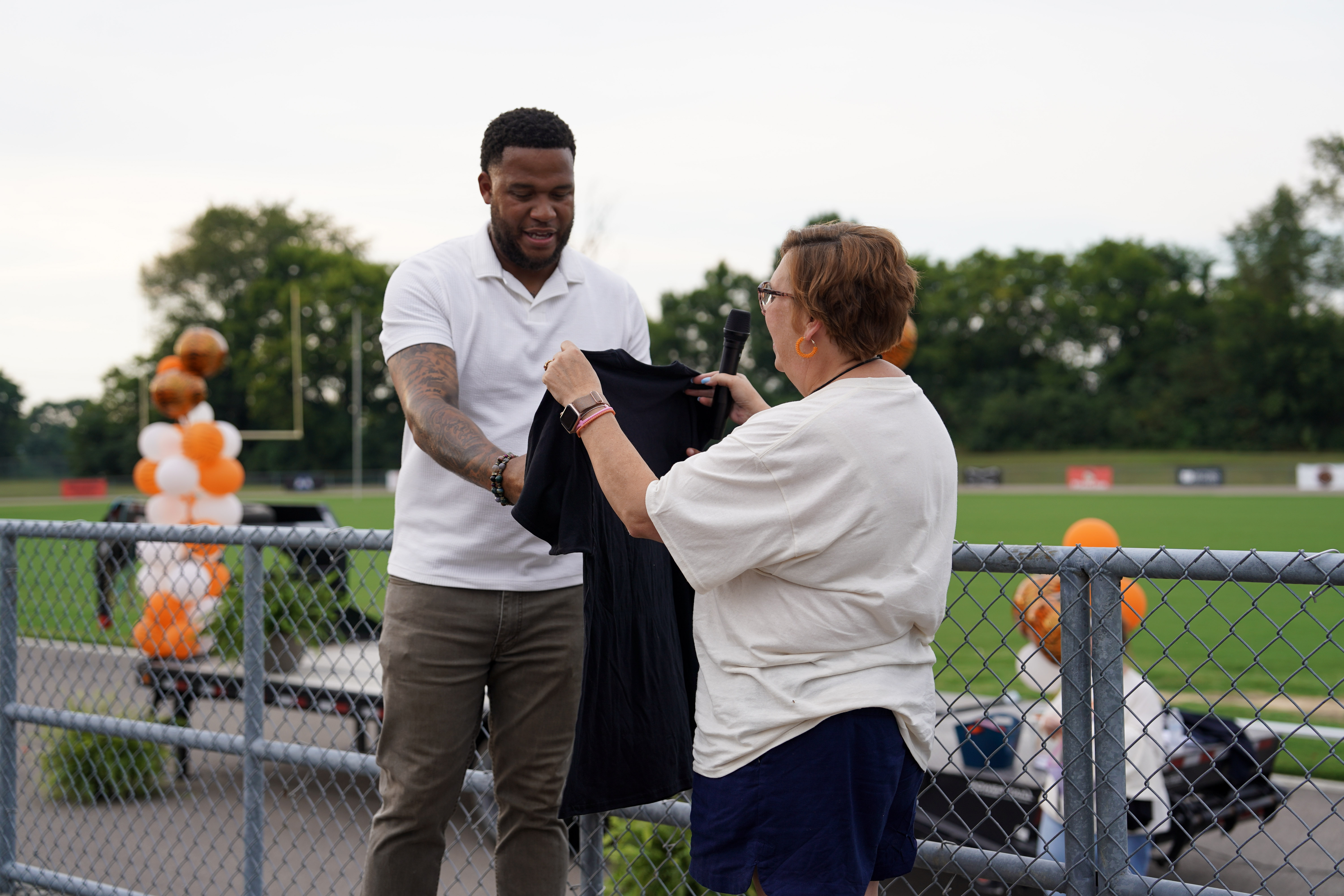 A man in a white polo hands a black T-shirt to a woman in a white shirt at an outdoor event near a sports field, with balloons and trees visible in the background.