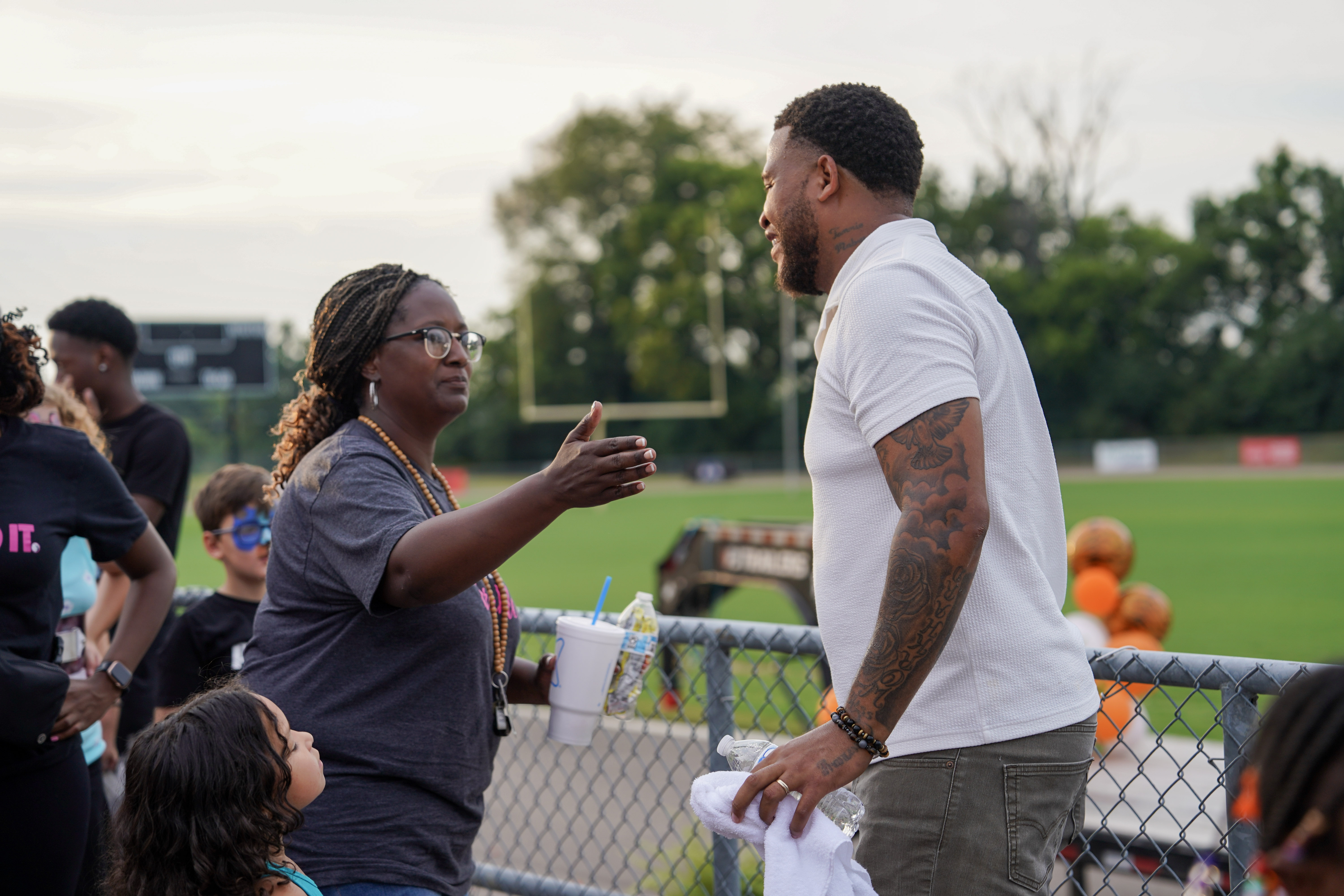 A woman with braids and glasses reaches out to greet a smiling man with tattoos at a sports field, while a young girl stands nearby. The background shows a green field and trees.