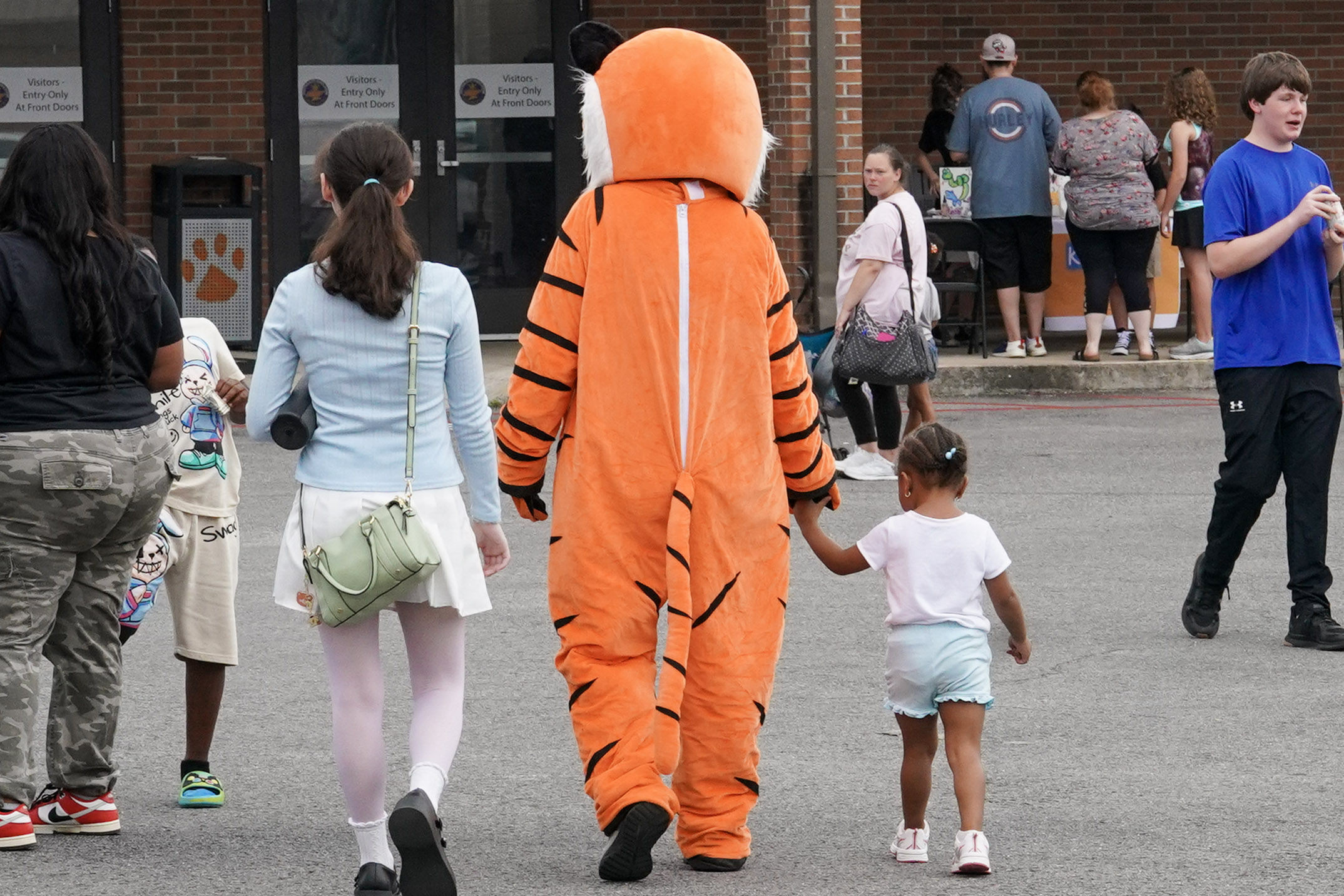 A person in a tiger costume walks outside holding hands with a young girl, while other people gather nearby in front of a building entrance.