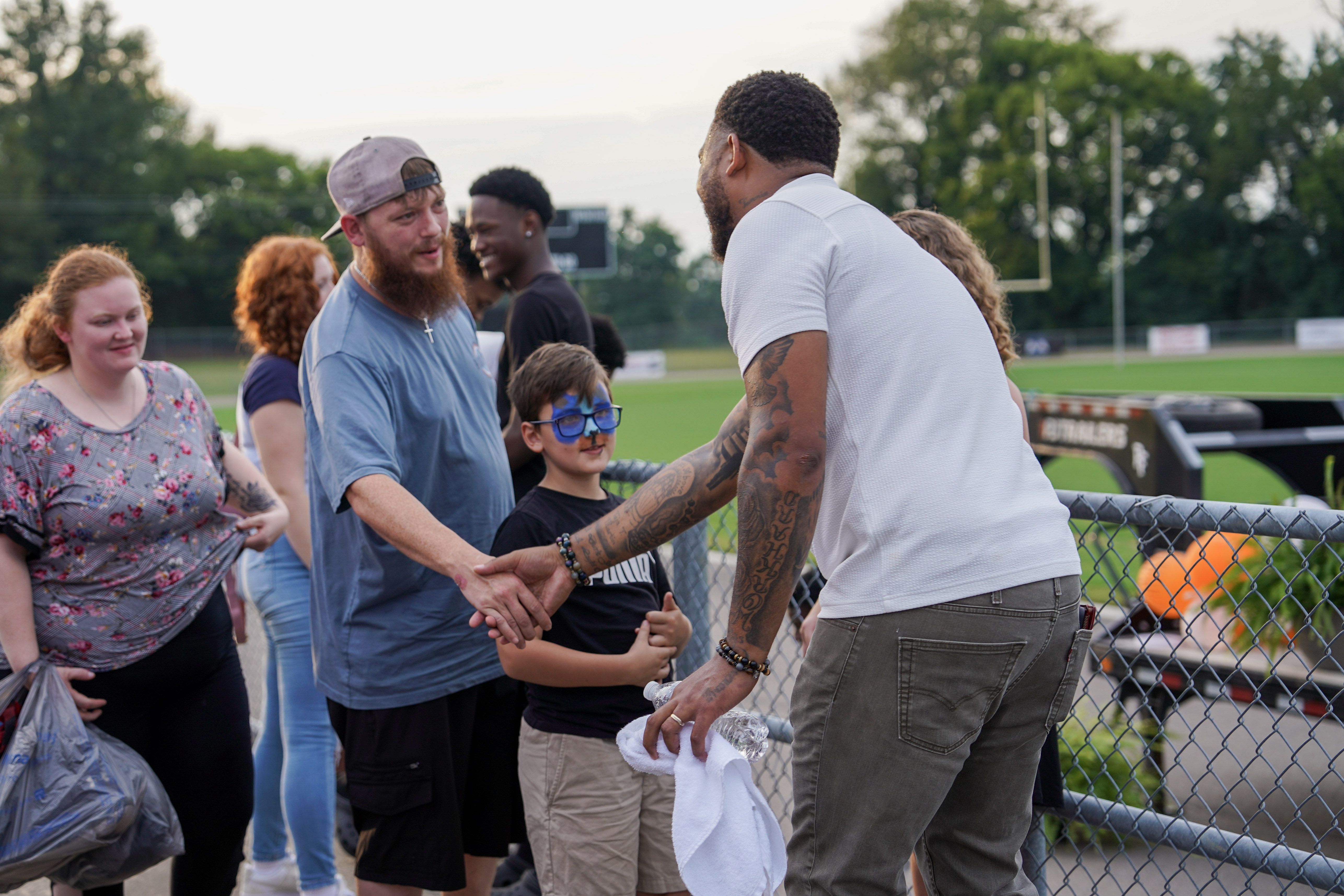 A man in a white shirt shakes hands with a bearded man by a fence at a sports field, as a smiling boy with blue glasses and several other people look on. Trees and a football field are visible in the background.