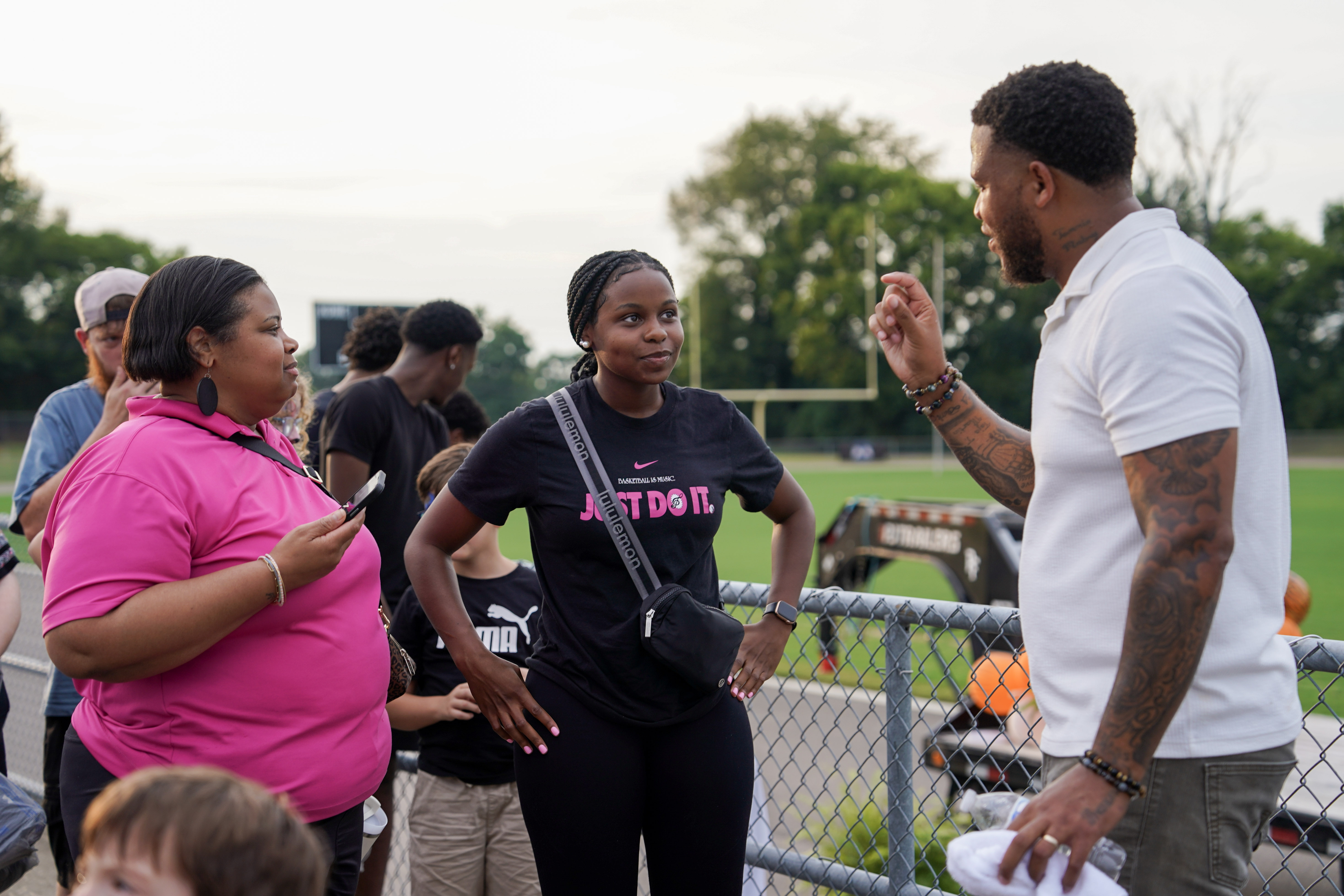 Three adults stand and talk by a metal fence at a sports field. One woman in a pink shirt holds a phone, another woman in black listens, and a man in a white shirt gestures as he speaks. People and trees are in the background.
