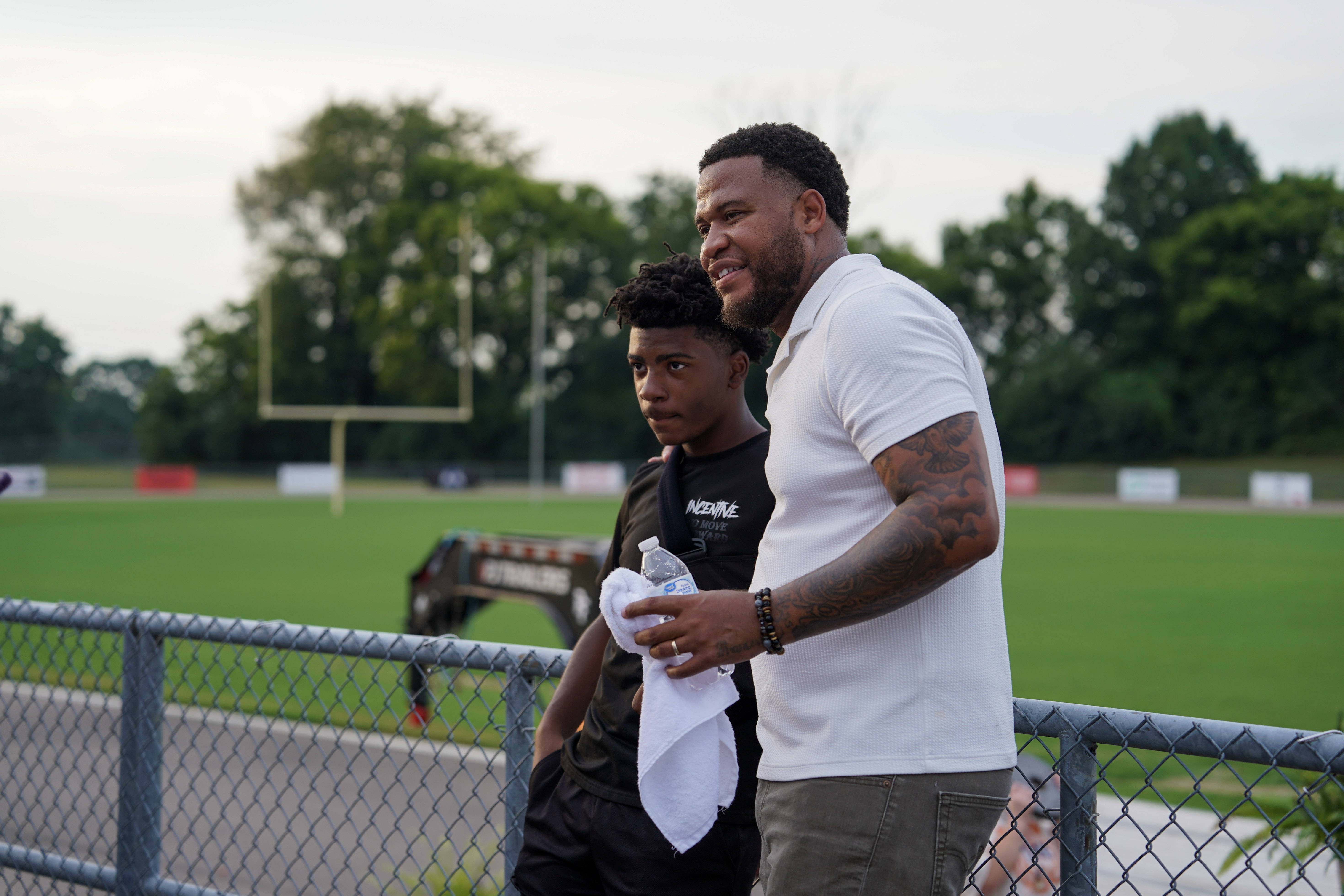Two people stand by a fence at a sports field, one in a black athletic outfit holding a towel, and the other in a white shirt smiling, with trees and a goalpost in the background.
