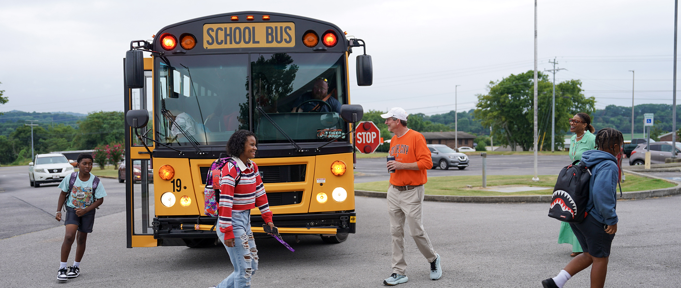 man in orange shirt and white cap greets students getting off of a school bus