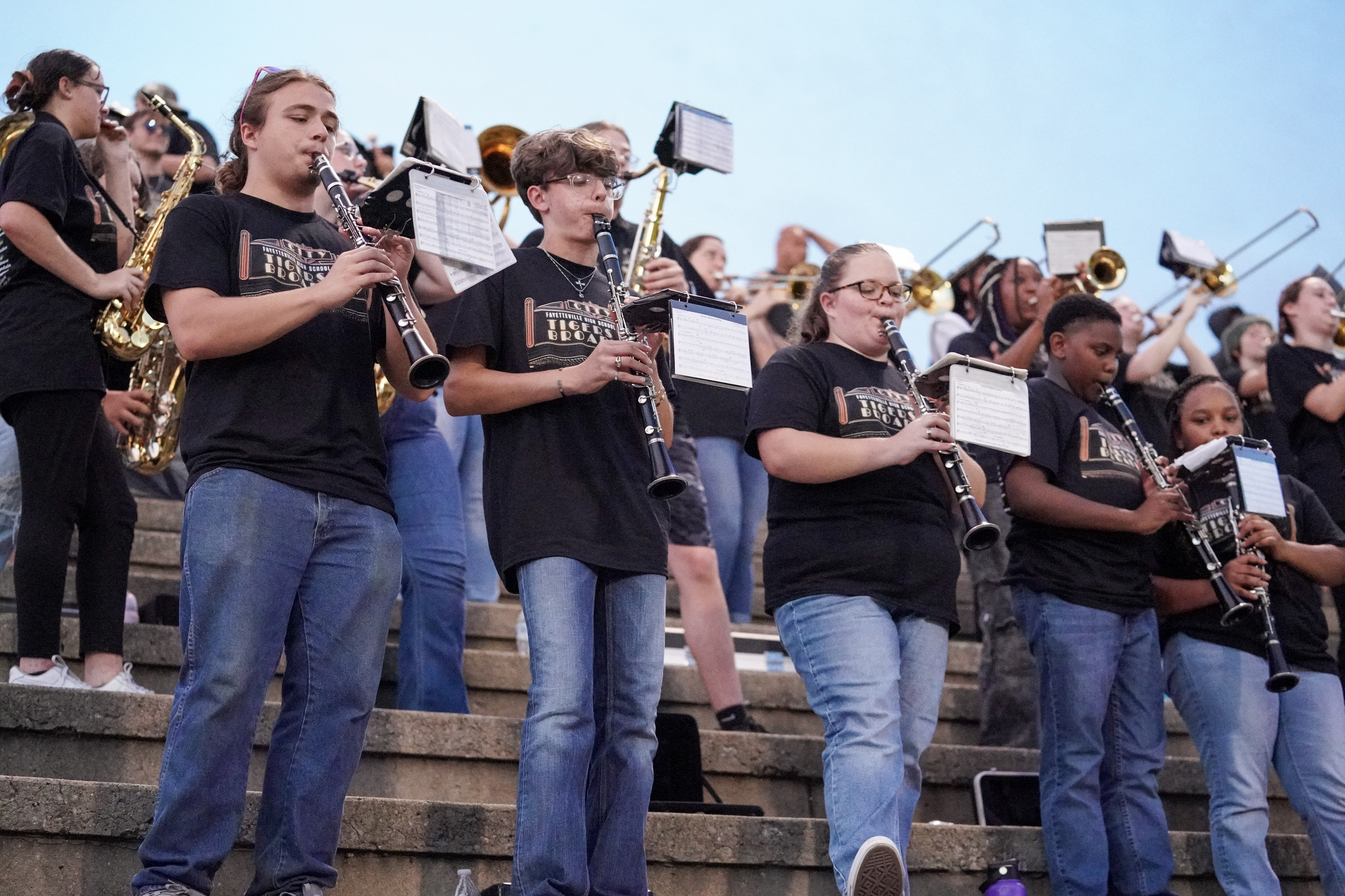 A group of students in matching black shirts and jeans play clarinets and other band instruments while standing on outdoor stadium steps, with more band members visible in the background.