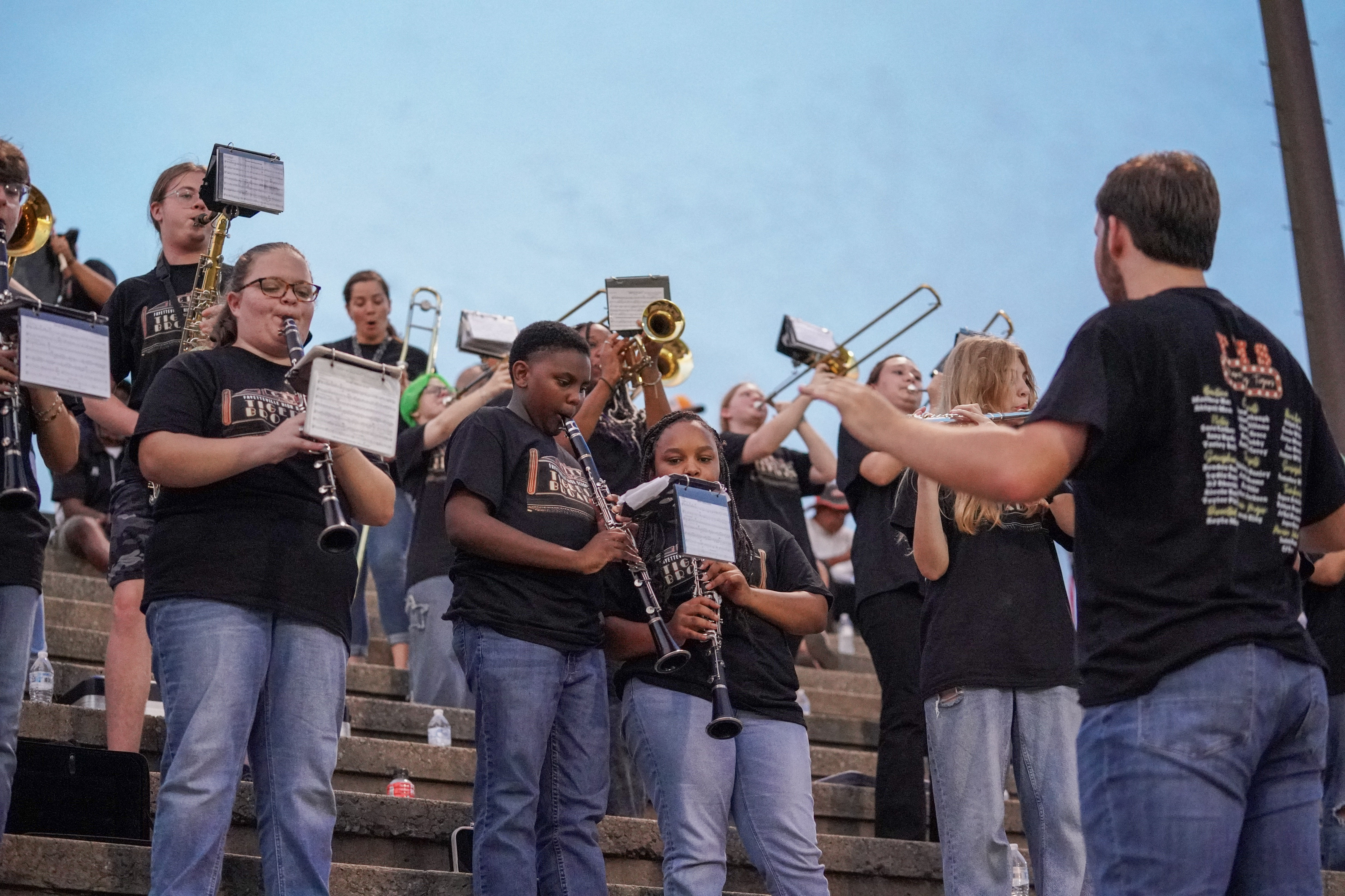 A marching band dressed in black shirts and jeans plays instruments on outdoor steps, with a conductor in front of them. The sky is clear and blue in the background.