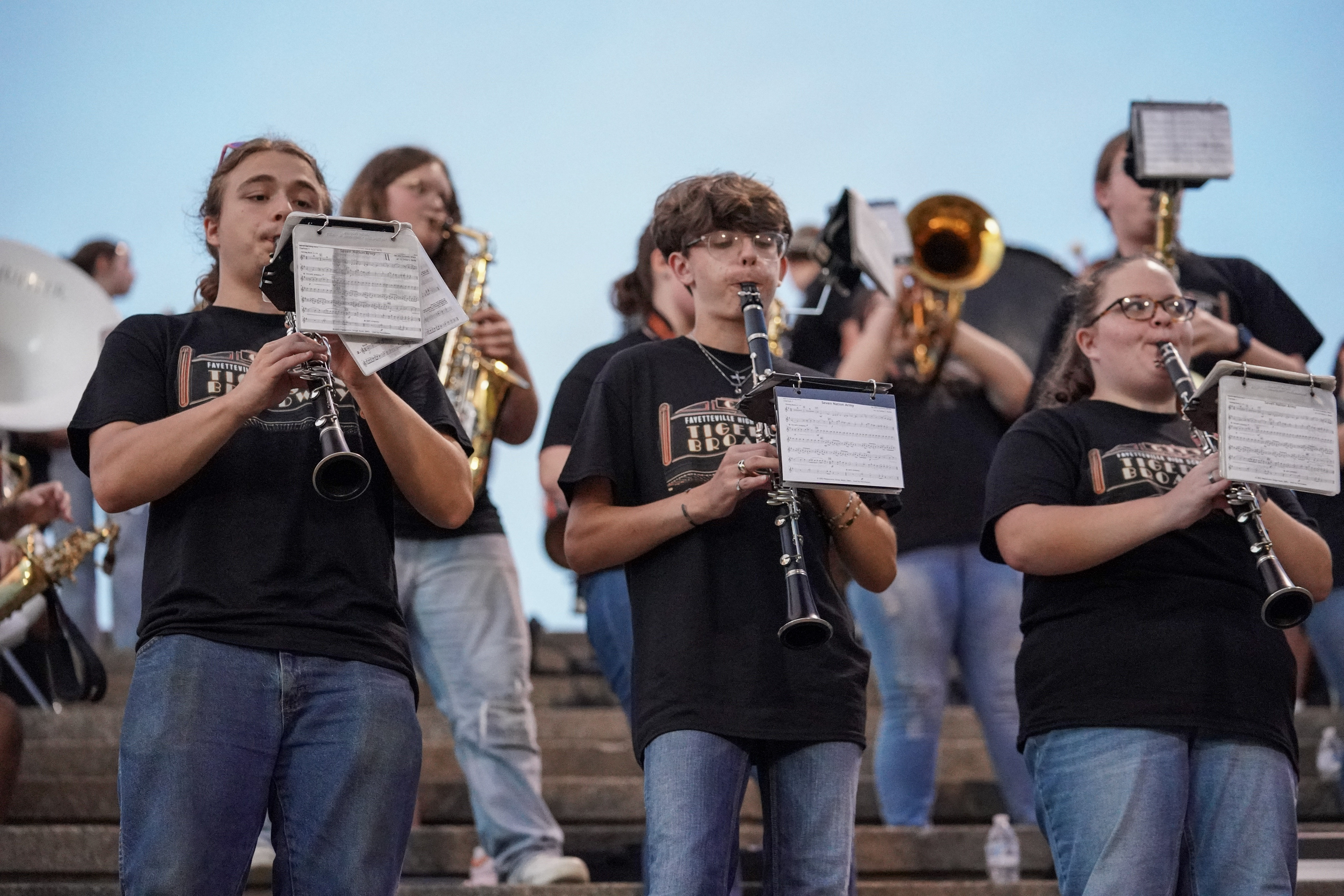 A group of marching band students in black T-shirts play clarinets and brass instruments while standing on stadium steps, with sheet music attached to their instruments. The sky is clear and blue.