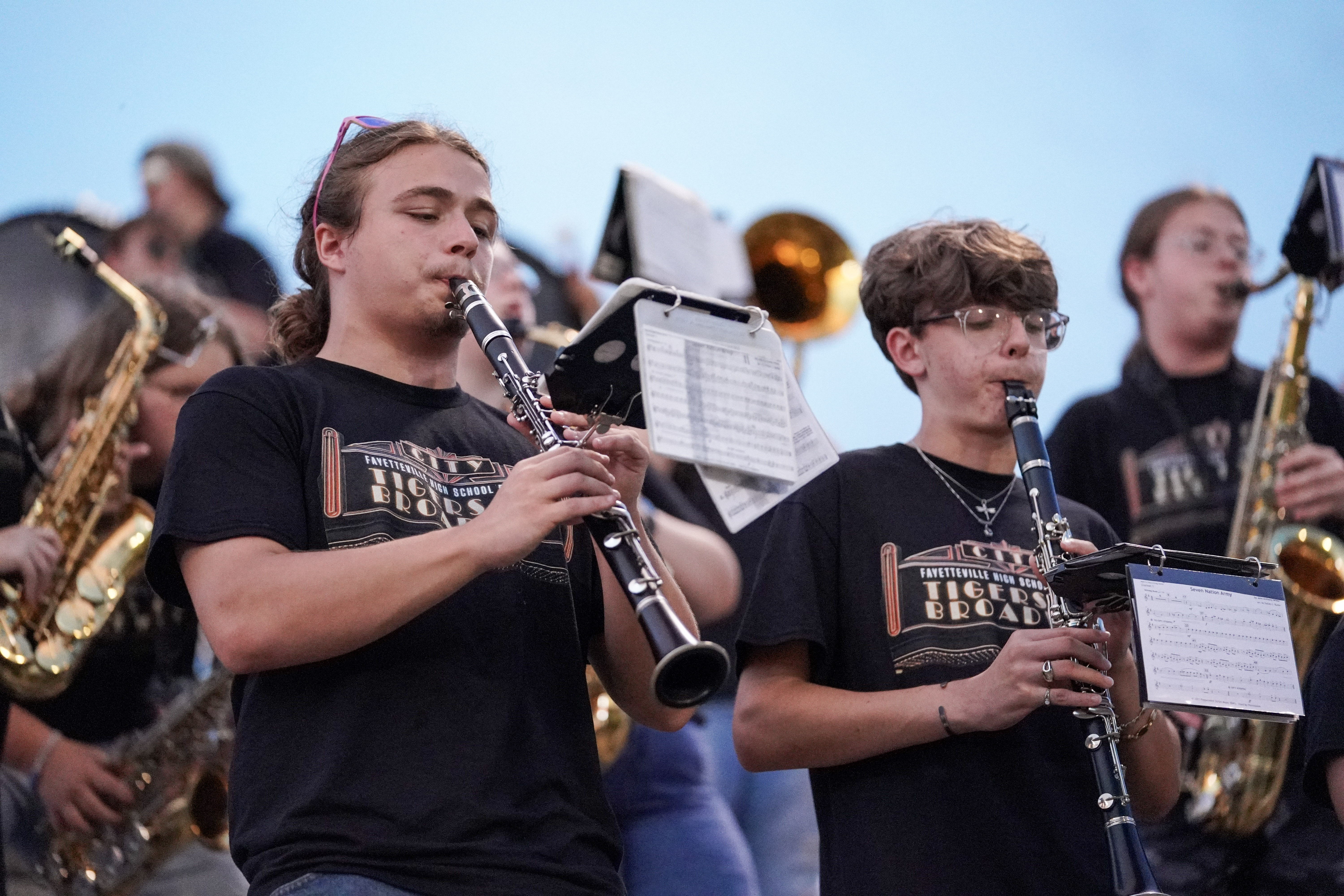 Two band members in black T-shirts play clarinets outside, surrounded by other musicians with brass instruments. Sheet music is clipped to their instruments, and the sky is clear in the background.