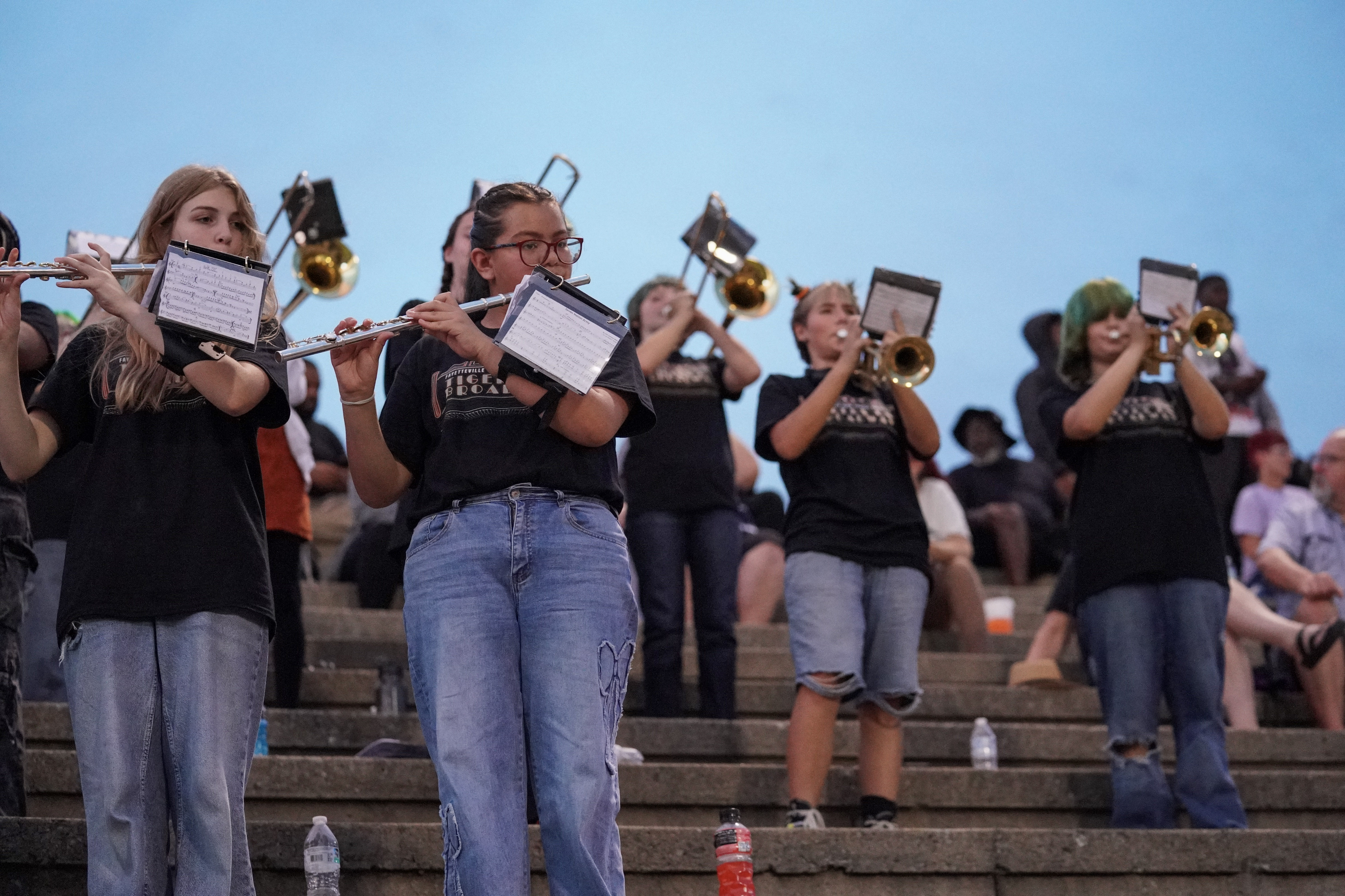 A group of students in black shirts and jeans play brass and woodwind instruments on outdoor steps, with people seated in the background under a clear blue sky.