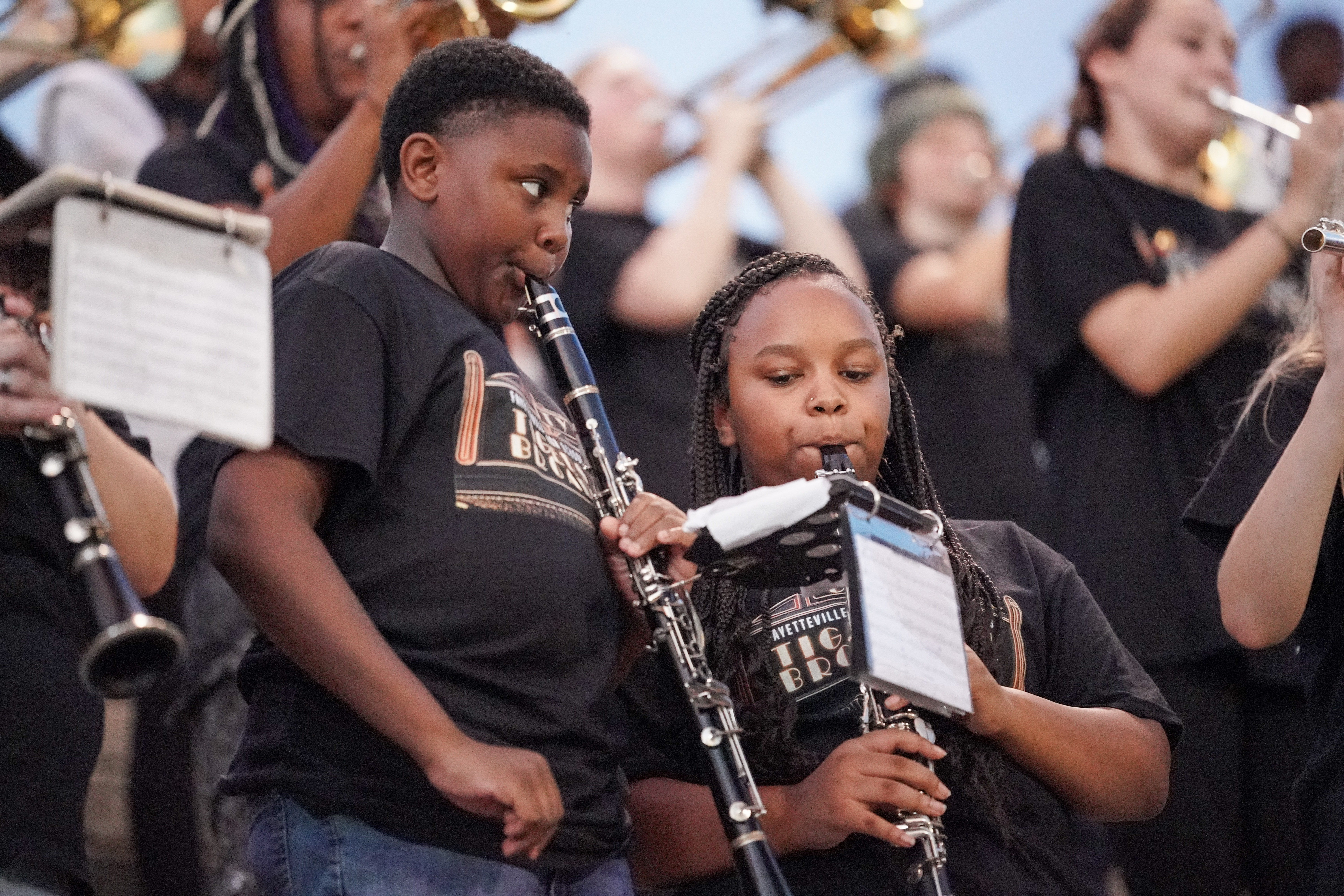 Two children wearing black band shirts play clarinets in a marching band, surrounded by other young musicians. Both are focused on their music, holding their instruments and reading from lyre-mounted sheet music.