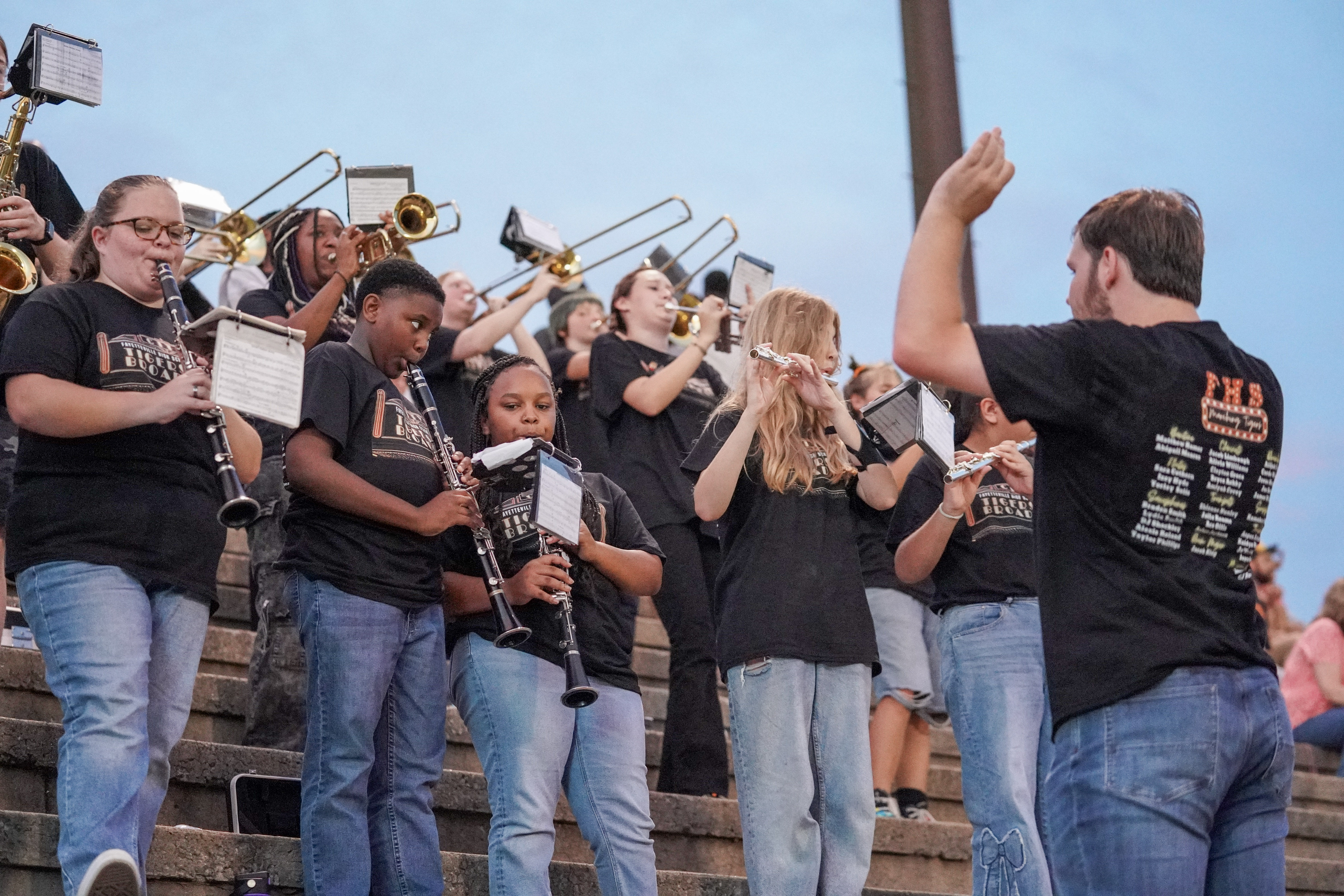 A band director leads a group of students playing instruments on outdoor bleachers. The students wear matching black T-shirts and play various brass and woodwind instruments. The sky is clear and blue.